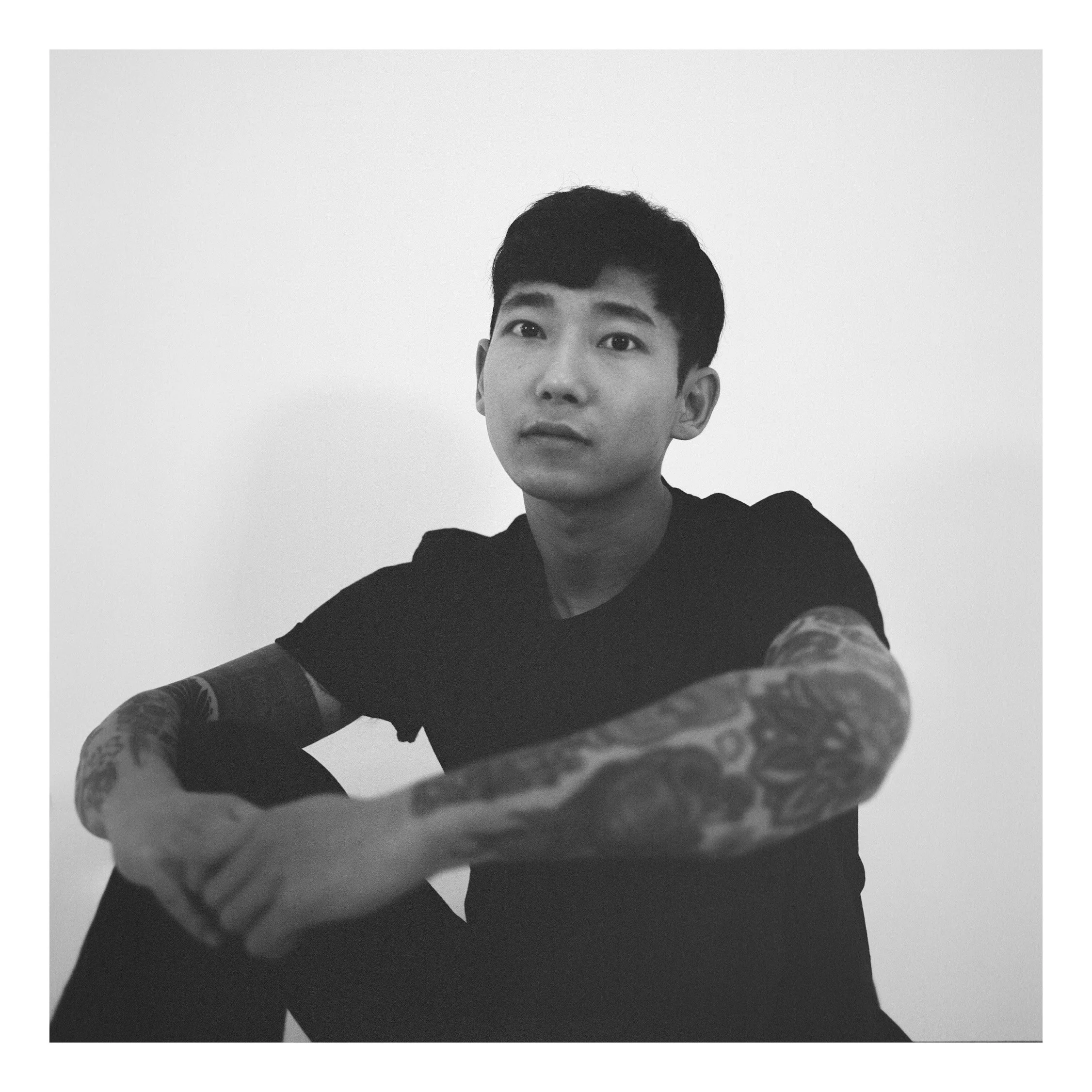 Black and white photo of a young man with short dark hair, looking at the camera, wearing a black t-shirt, with tattoos on both arms, sitting against a plain white background.