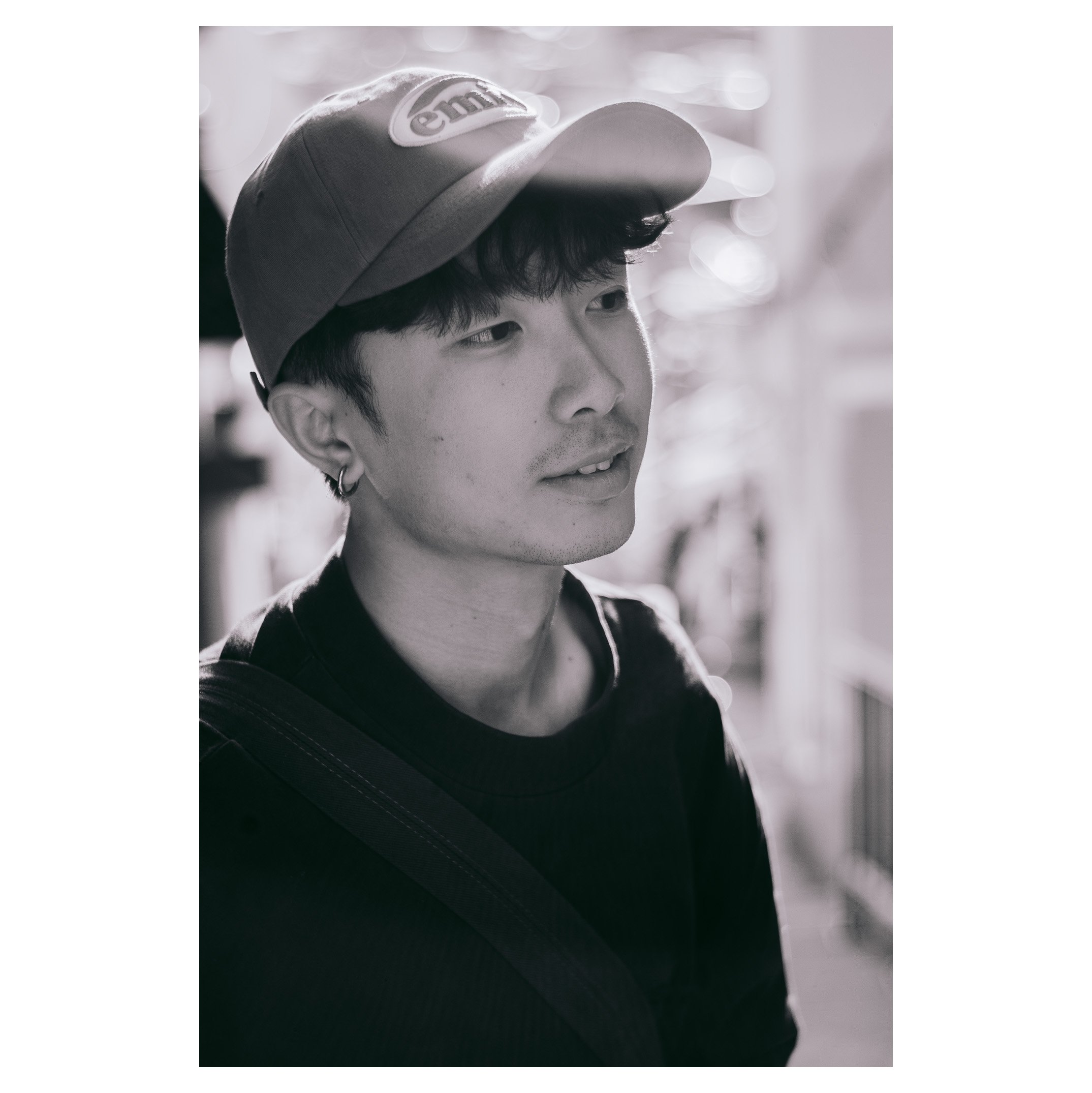 A young man with dark hair wearing a baseball cap, earrings, and a black shirt, looking slightly to the side with a thoughtful expression.