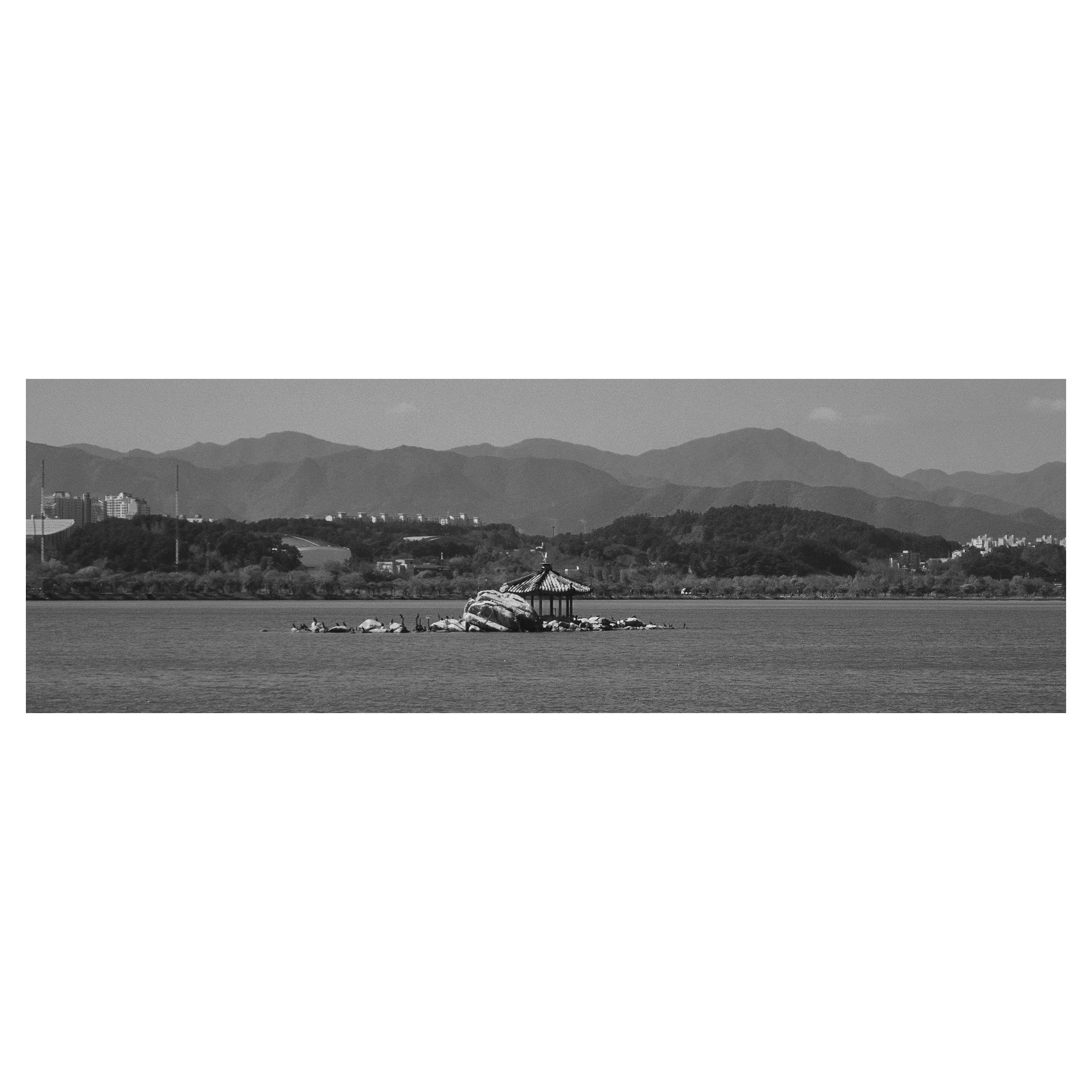 A black-and-white photo of a small pavilion on a rock island in a lake, with mountains and buildings in the background.