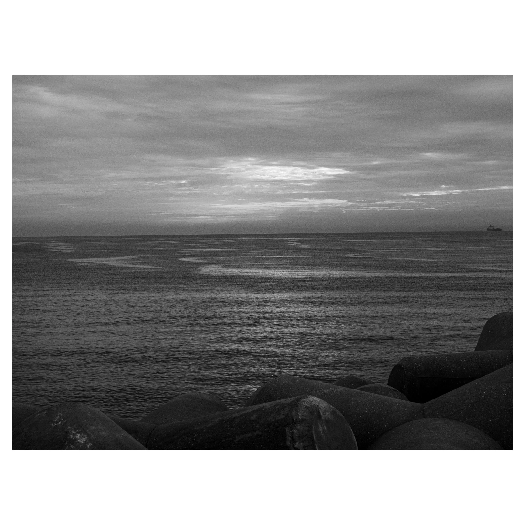 Black and white photograph of a calm ocean with a cloudy sky, large rocks along the shoreline, and a ship on the horizon.
