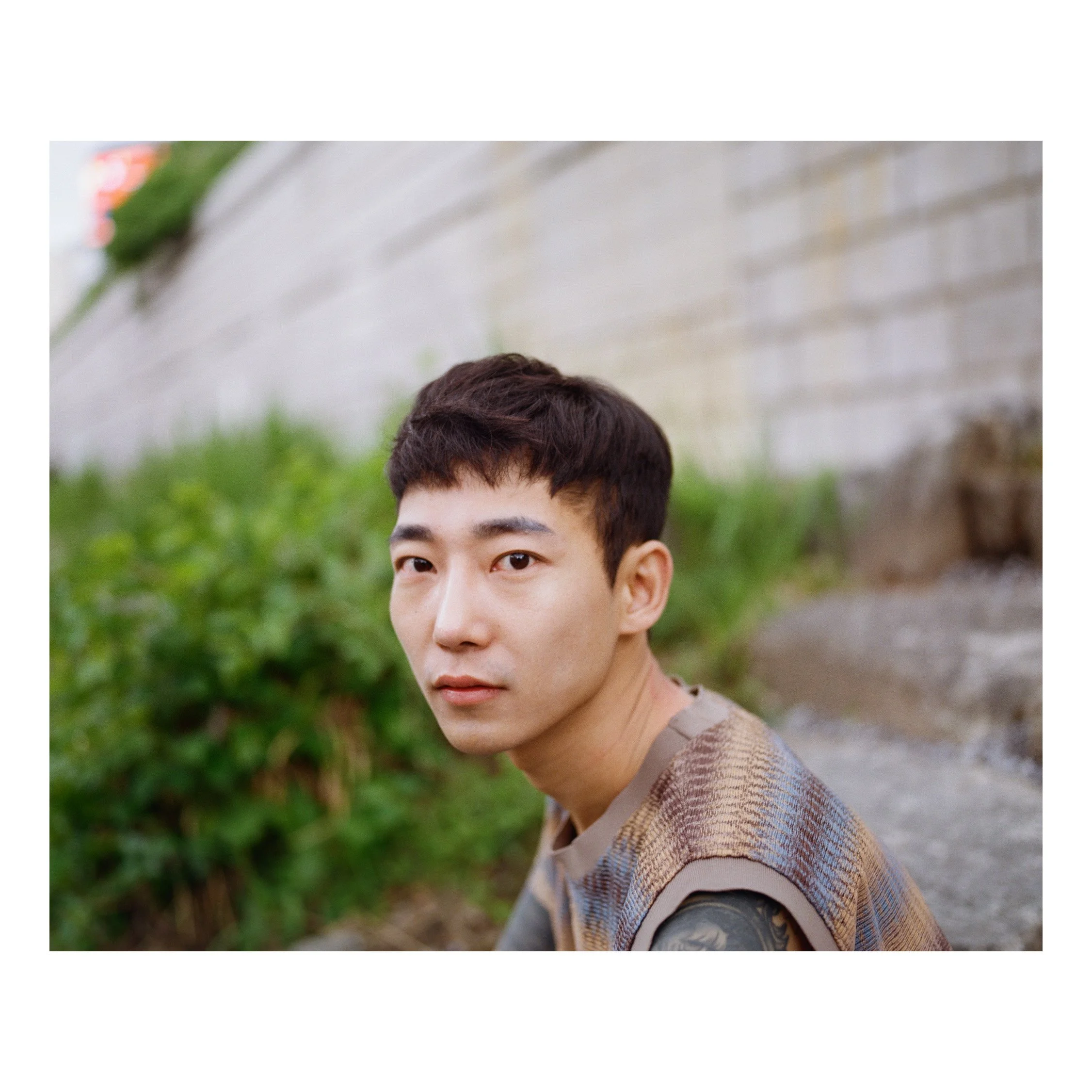 Portrait of a young man with short dark hair, sitting outside near a concrete wall, with green bushes in the background.