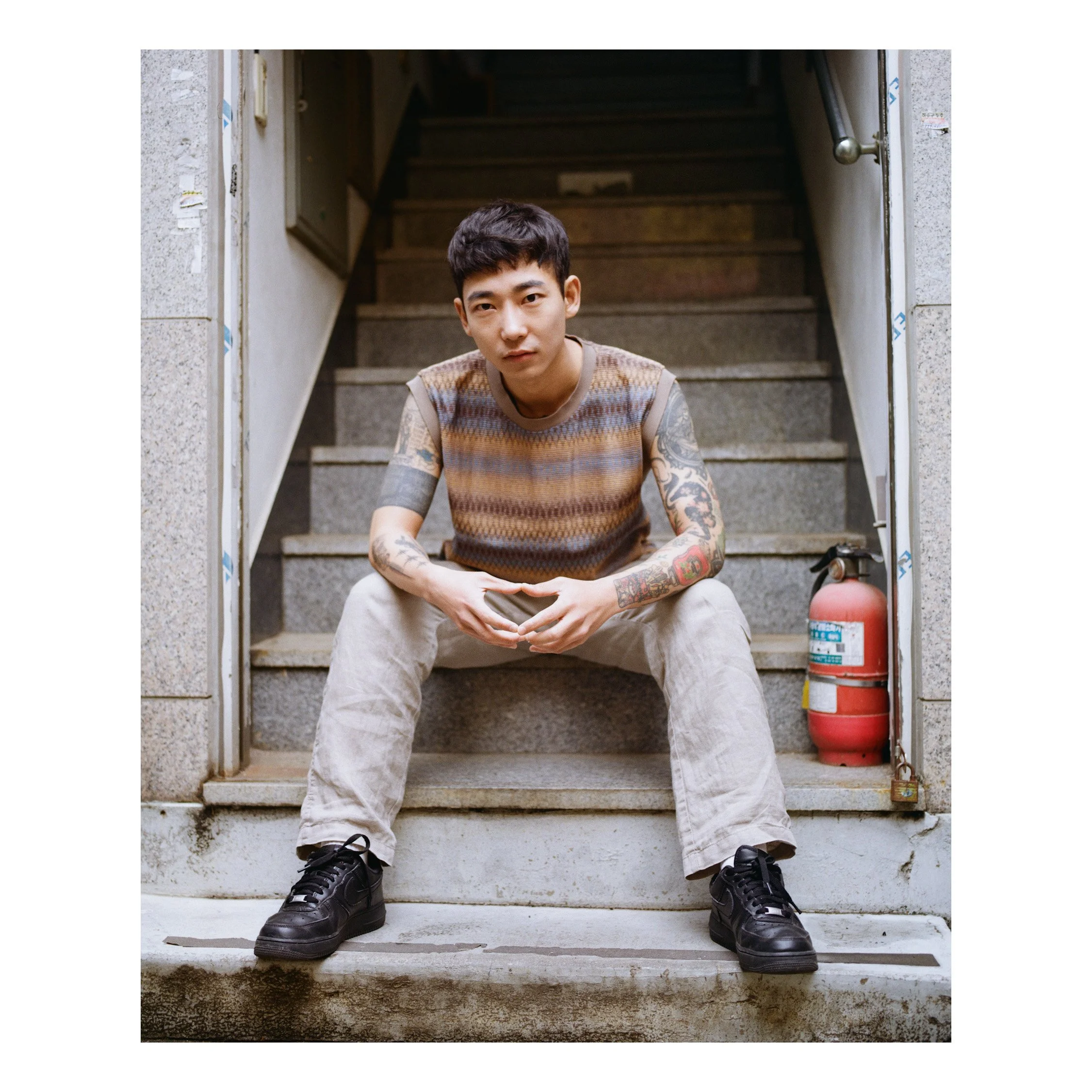 A young man with short dark hair, sitting on stairs in an urban setting, wearing a multicolored patterned sleeveless shirt, beige pants, and black sneakers, with tattooed arms, holding a smartphone, and a fire extinguisher is visible beside him.