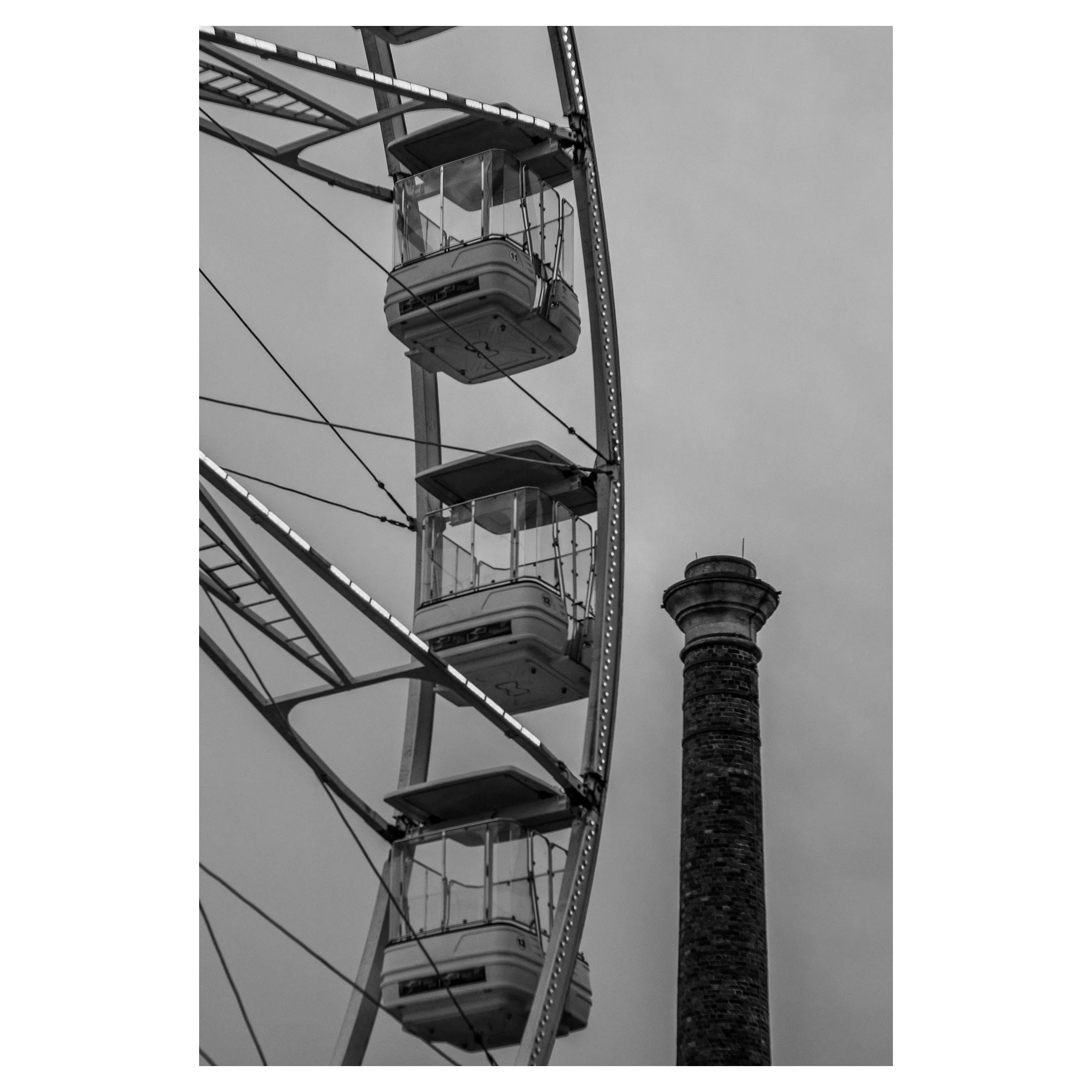 Black and white photo of a Ferris wheel with enclosed gondolas and a tall brick chimney in the background.