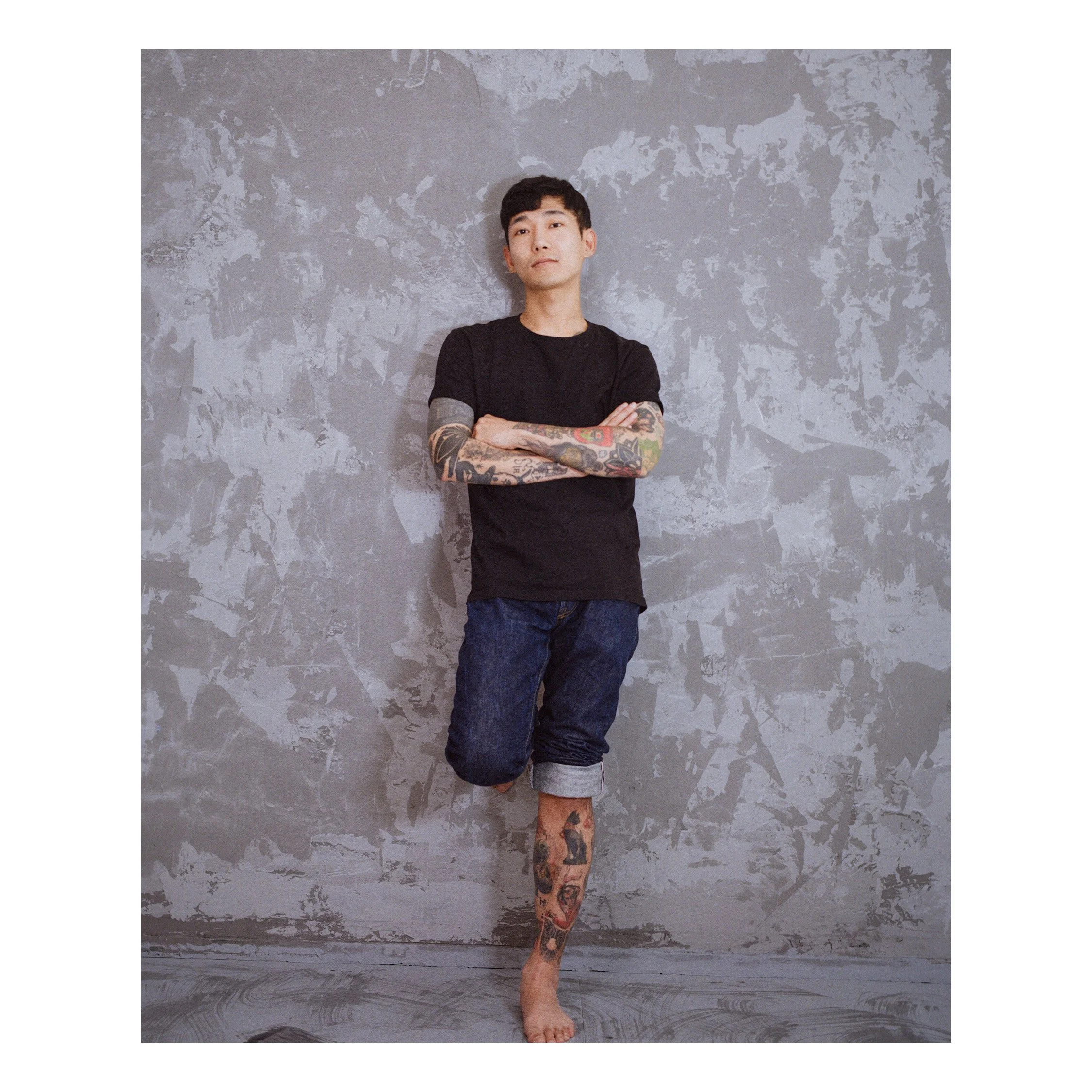 A young man with tattoos standing against a textured gray wall. He is wearing a black t-shirt and rolled-up denim shorts, with arms crossed and one leg bent, barefoot.