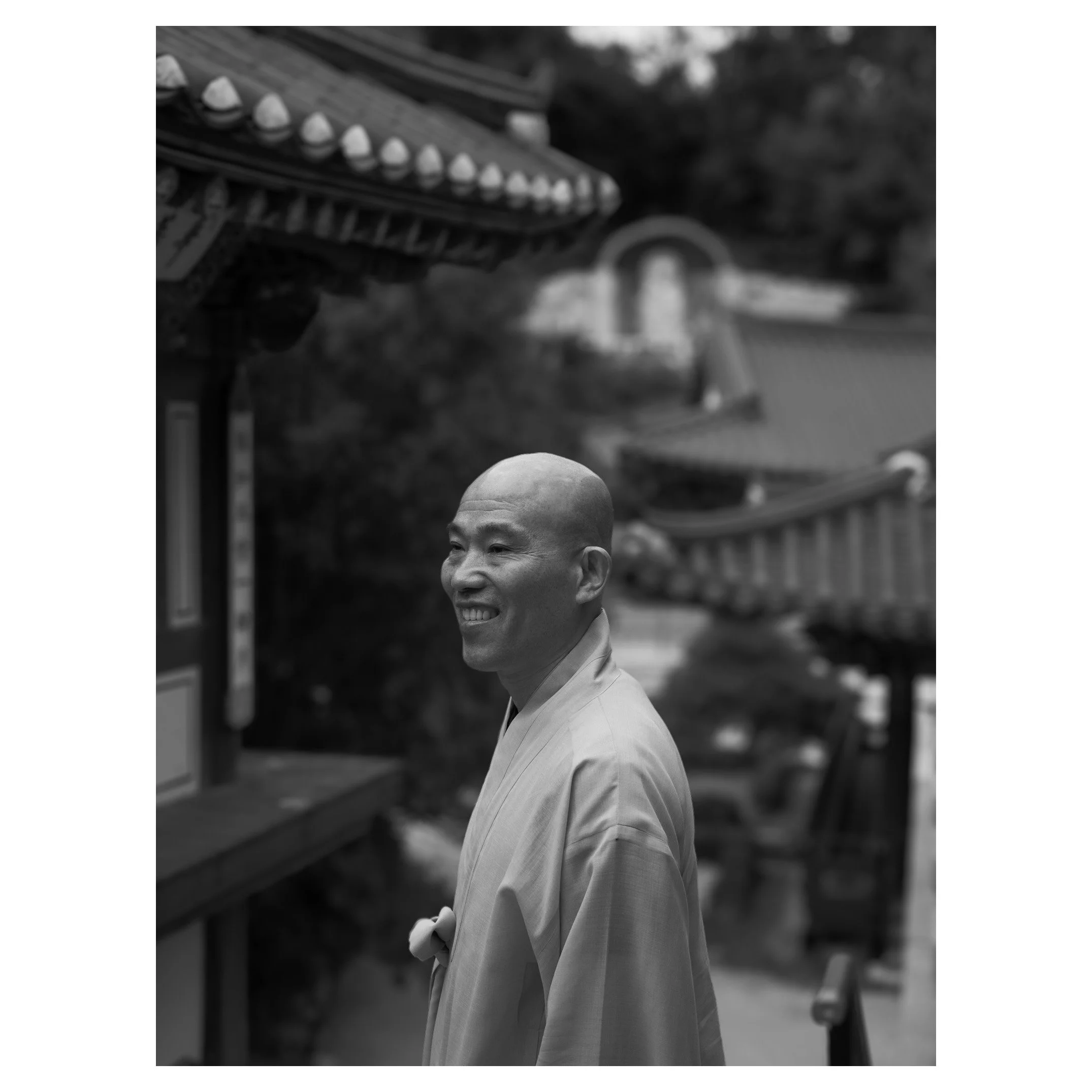 A Buddhist monk wearing traditional robes smiling outdoors near temples with curved roofs and decorative elements.