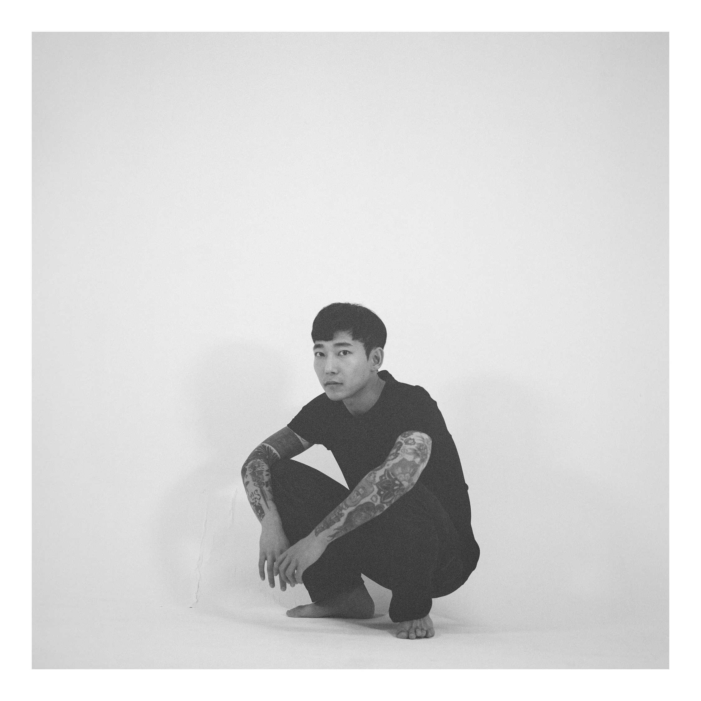 A young man with tattoos wearing a black t-shirt and black pants, squatting barefoot against a plain white background.