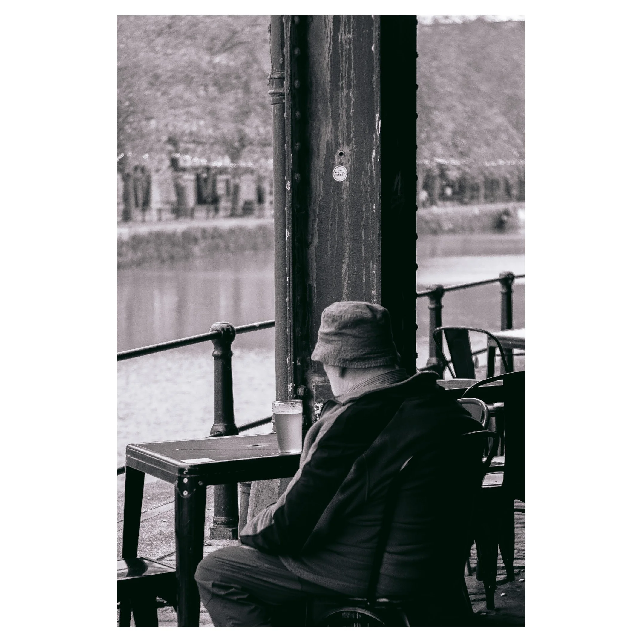 A person sitting alone at a table outside by a river, wearing a bucket hat and a jacket, with a pint of beer on the table.