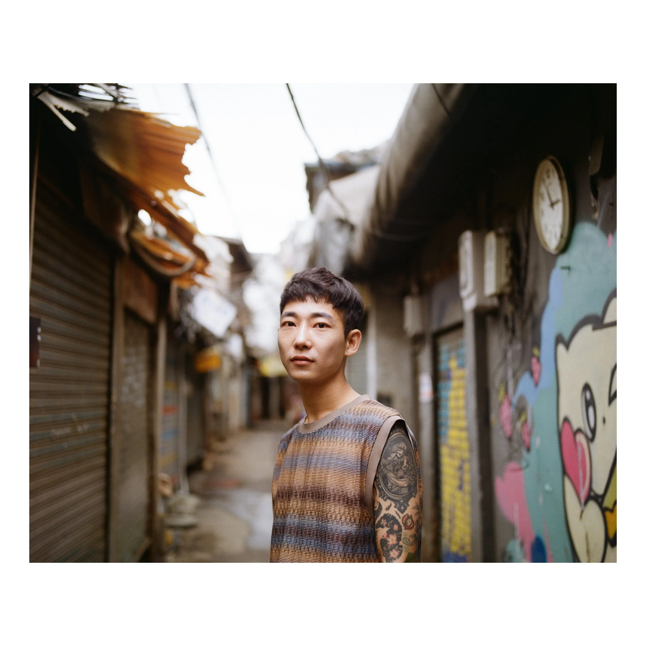 A young man standing in an urban alleyway with graffiti on the wall, wearing a sleeveless striped shirt, looking at the camera.