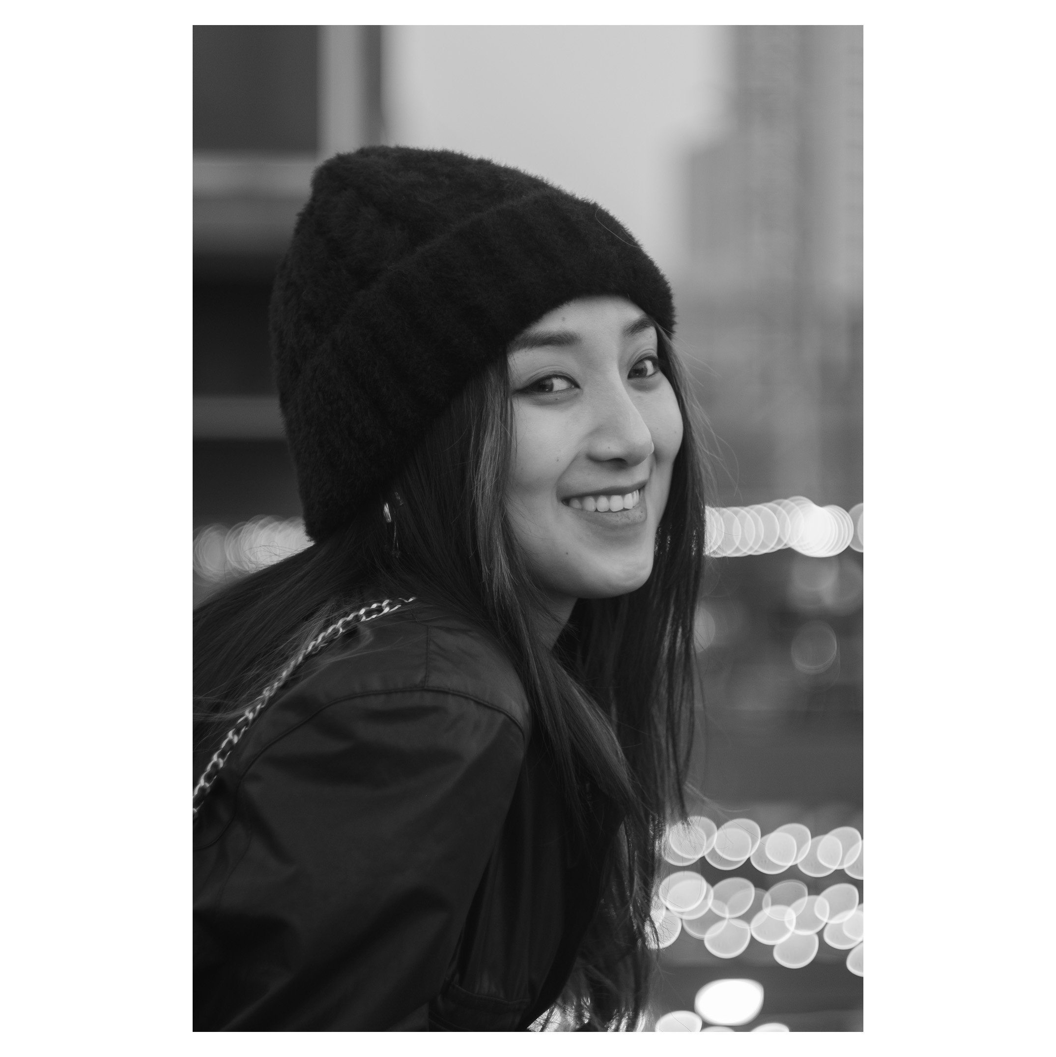 A smiling young woman with long dark hair wearing a beanie and a jacket, with city blurred lights in the background.