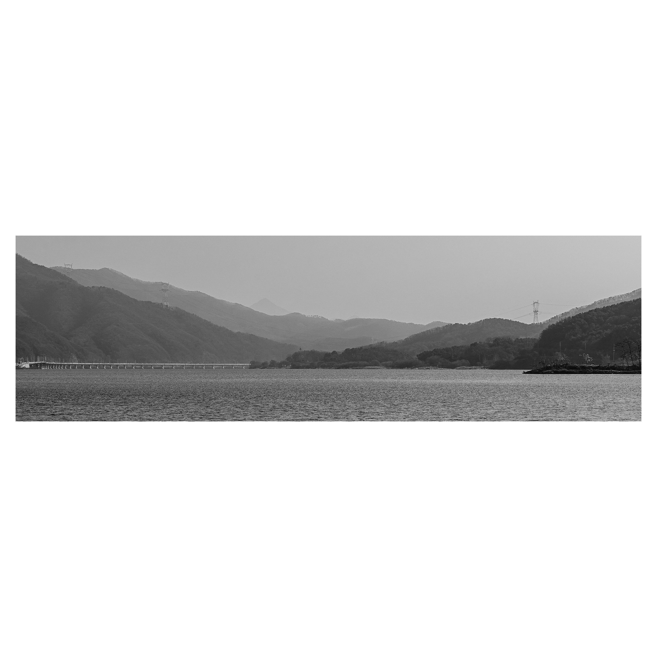 Black and white panoramic view of a lake with surrounding mountains and hills, with a bridge extending across the water on the left side.