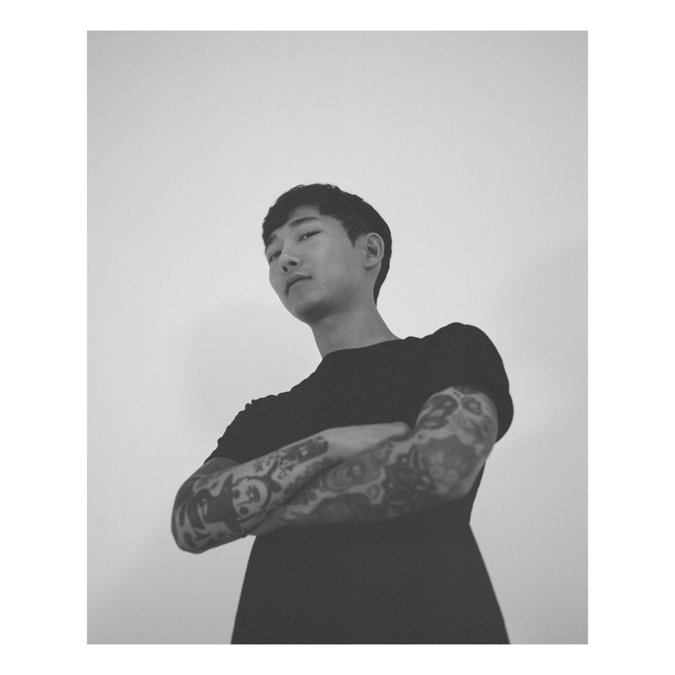 Black and white photo of a young man with short hair and tattoos on his arms, standing with arms crossed and looking slightly upward.