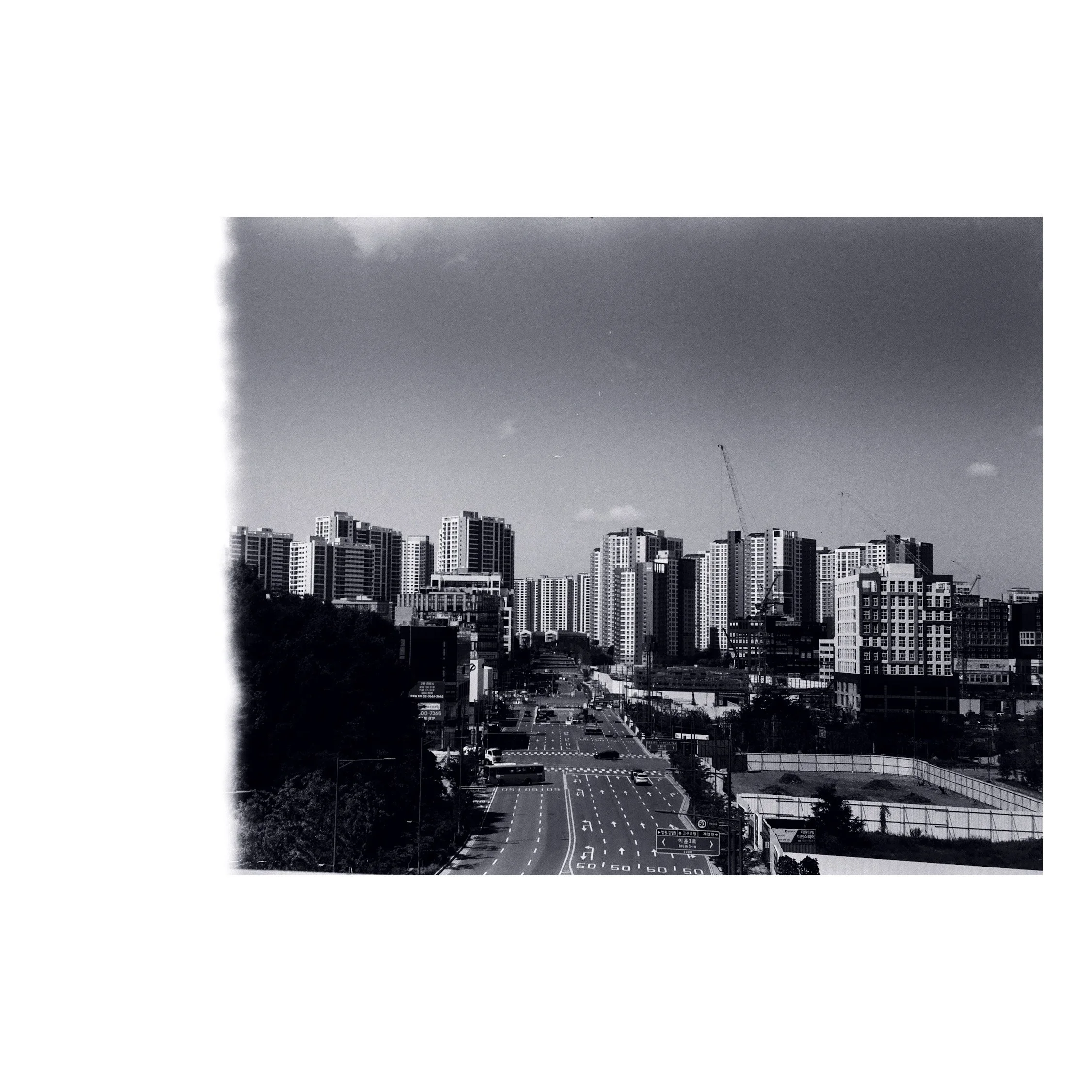 Black and white photograph of a cityscape with tall buildings, construction cranes, and a road in the foreground.