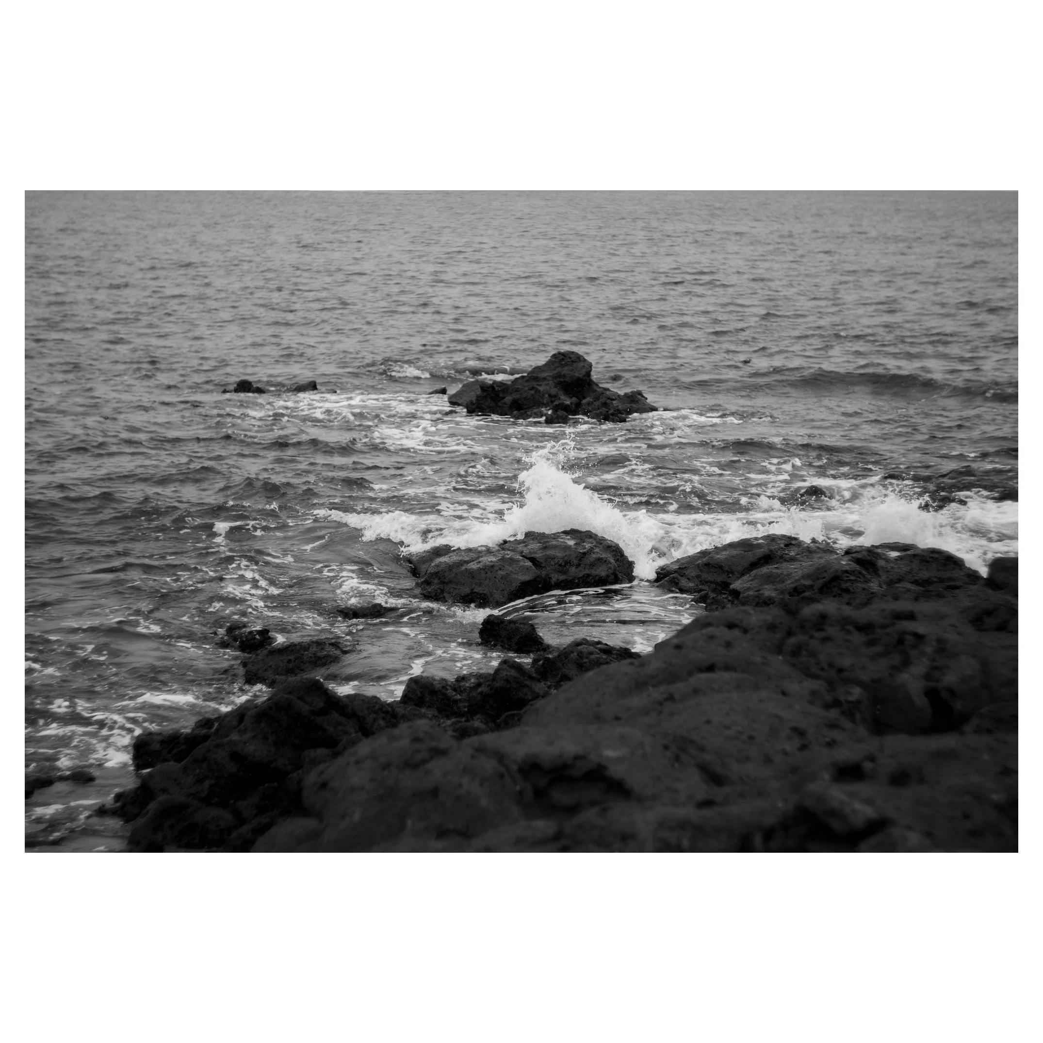 Black-and-white photo of rocks along a shoreline with waves crashing against them, extending into the ocean.
