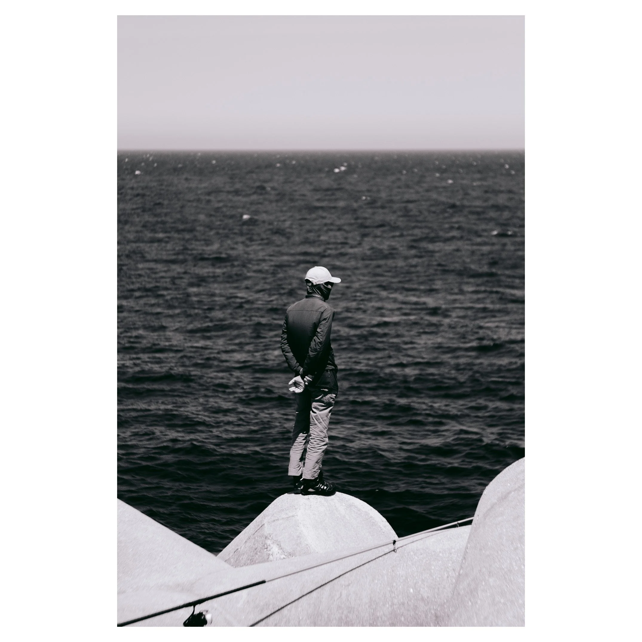 A person standing on a large rock by the water, wearing a cap, jacket, and pants, looking out at the ocean in black and white.
