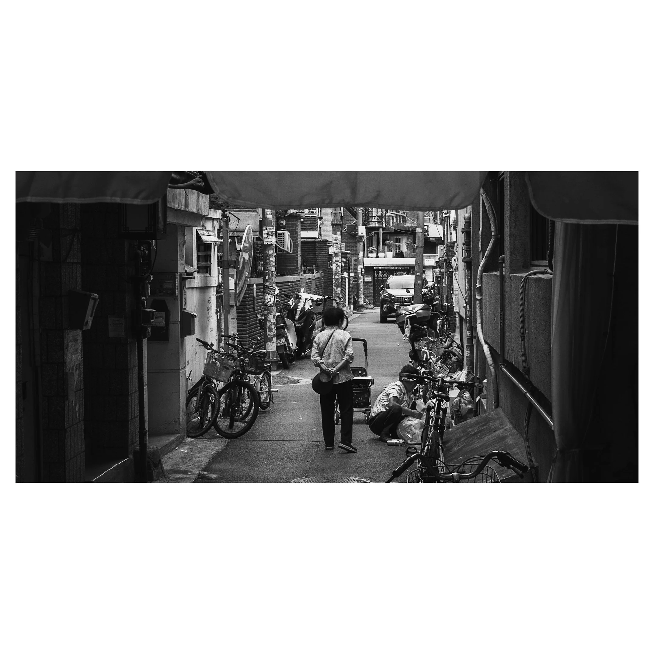 A narrow alley with bicycles and scooters parked along the sides, a woman carrying a hat walking away, and a person sitting on the ground fixing a bicycle, in an urban environment.