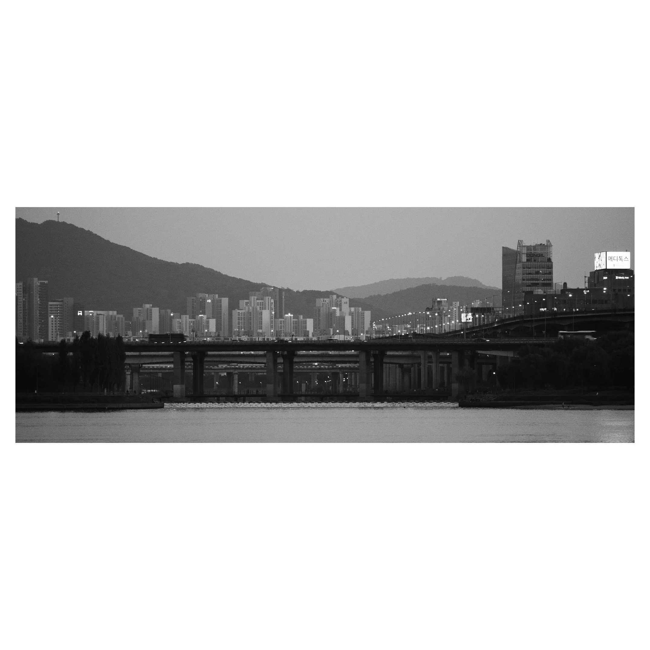 Black and white cityscape with high-rise buildings, a bridge over a river, and mountain in the background.