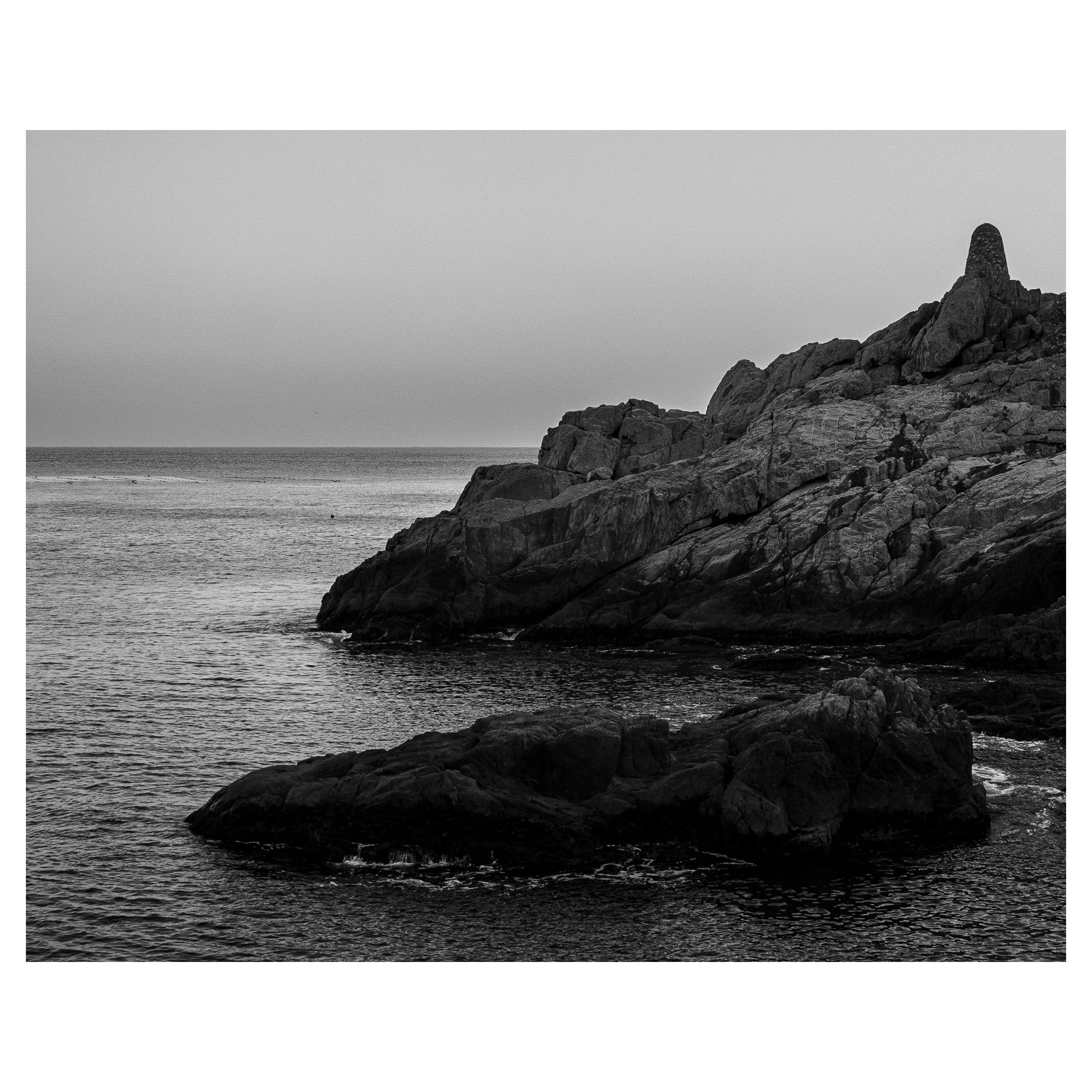 Black and white photograph of rocky coastline with large rocks extending into the ocean, calm water, and a distant horizon under an overcast sky.
