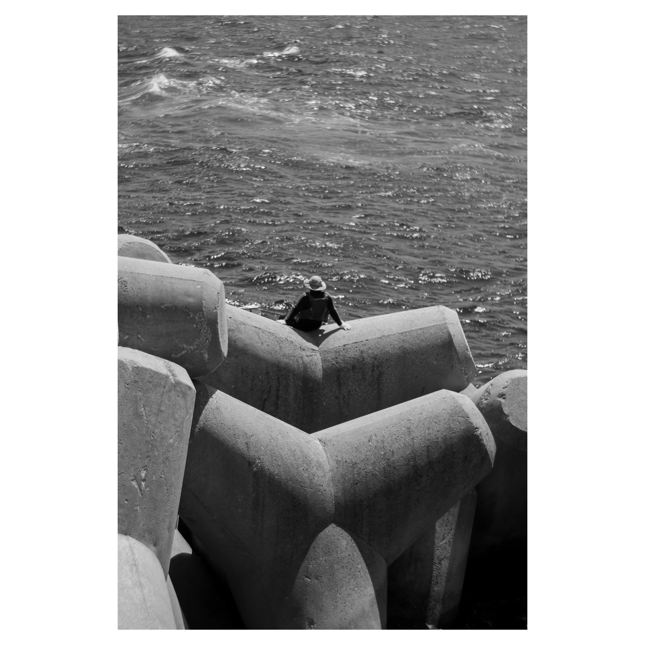 A person wearing a hat sitting on concrete breakwater blocks near the water, viewed from behind.