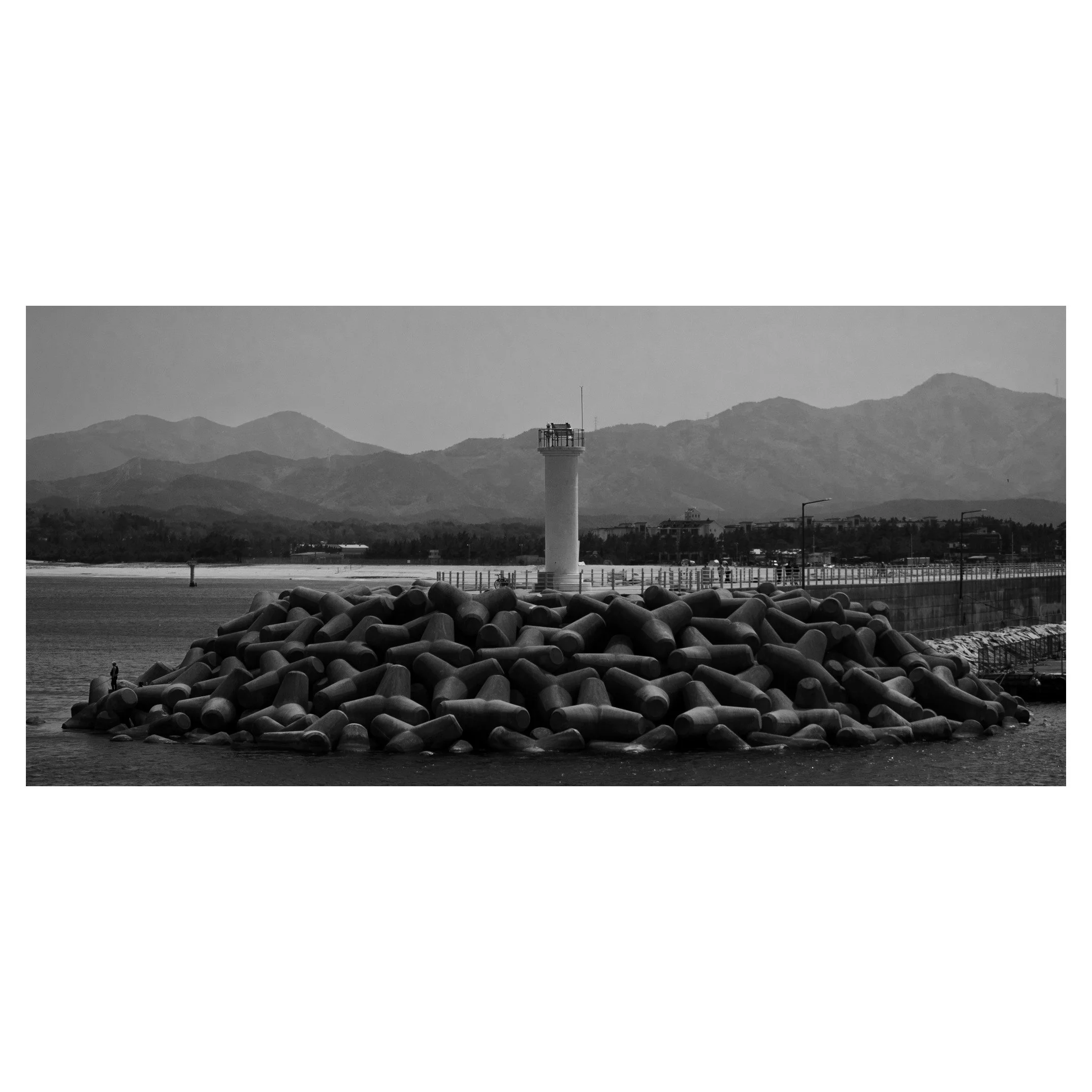Black and white photo of a lighthouse on a breakwater built with large concrete sea barriers, with mountains in the background.