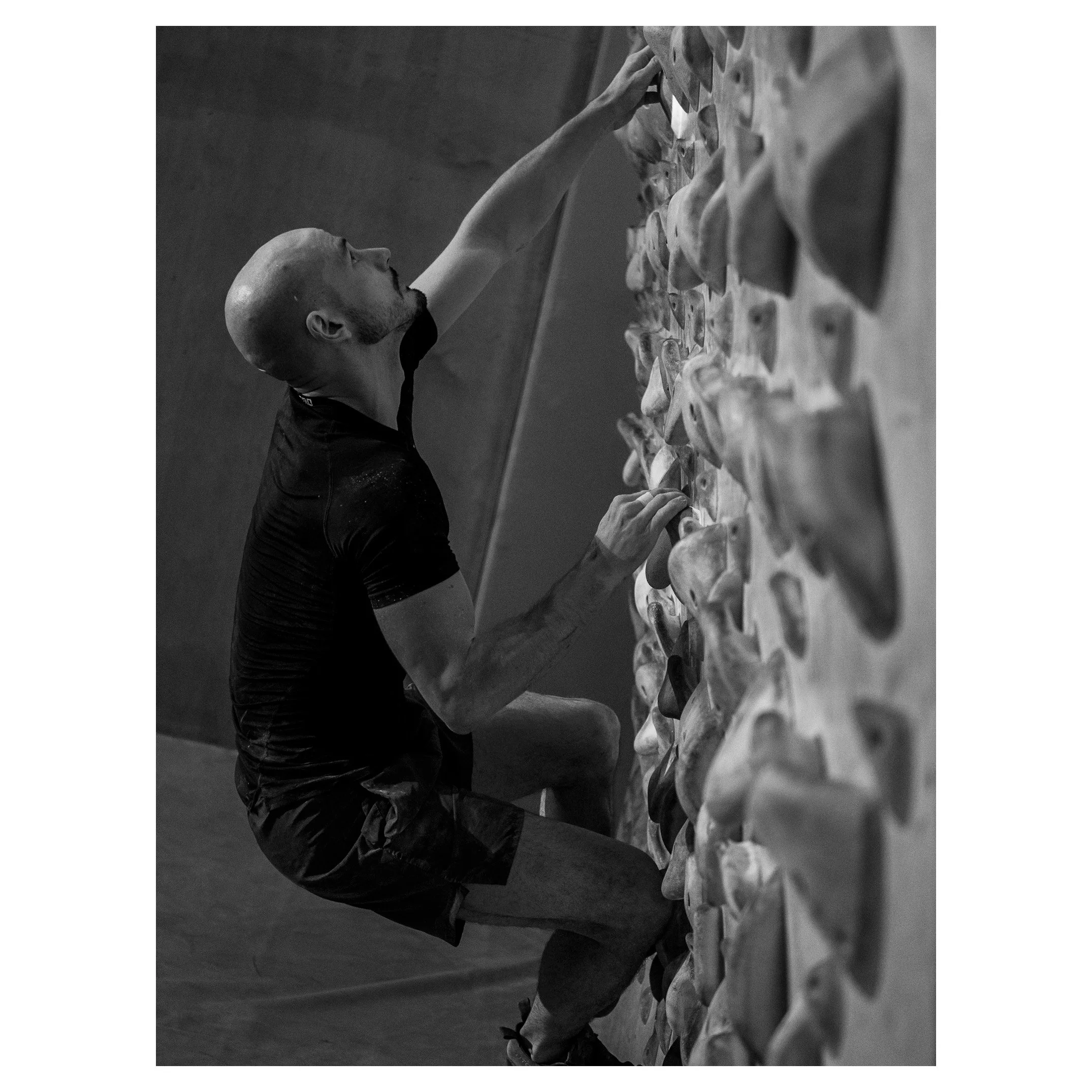 Man climbing an indoor rock wall, reaching for holds with his right hand, with a bald head and short beard, wearing a black shirt and shorts.
