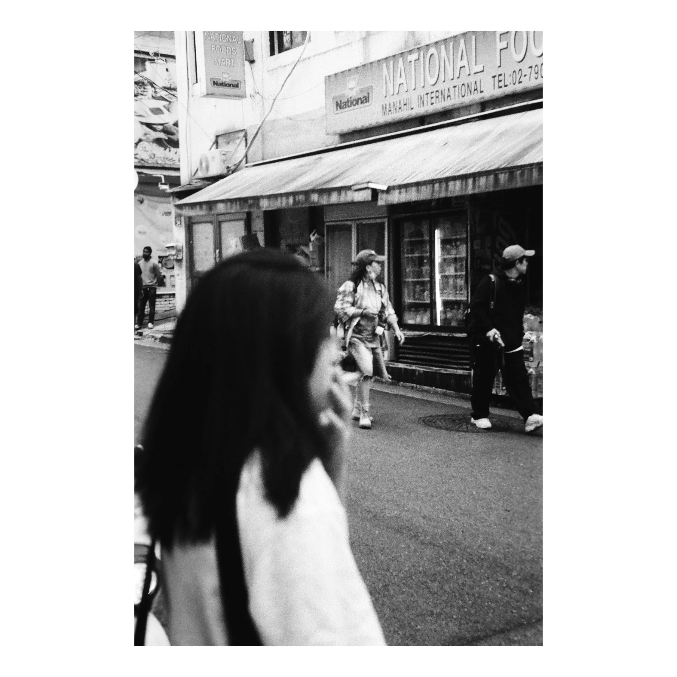 Black and white photo of an urban street scene with pedestrians walking past a store named 'National Food.' The store has a large awning and windows displaying products. The image includes a person in the foreground with long dark hair, out of focus,