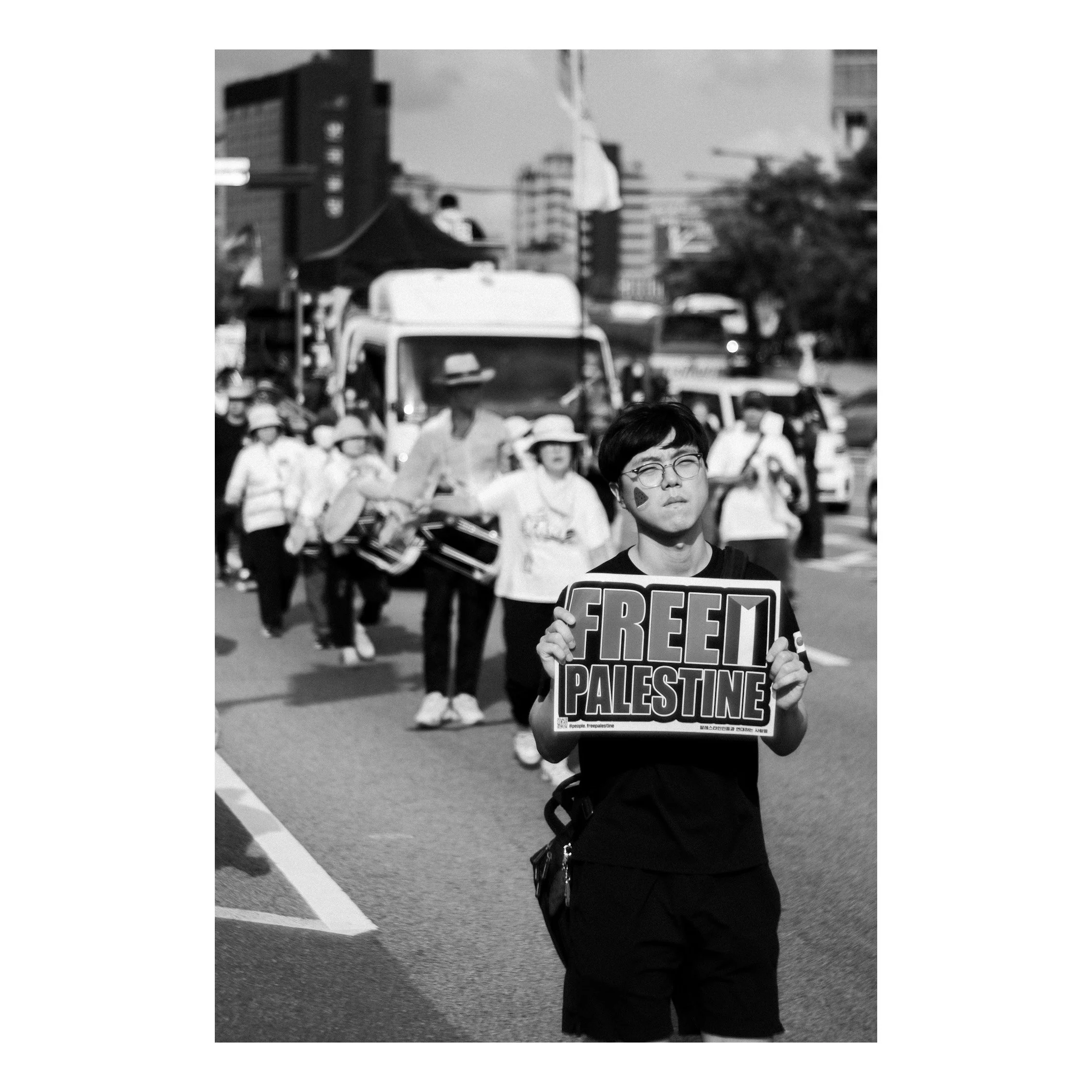 A young person holding a sign that says 'FREE PALESTINE' in a protest or demonstration, with a group of people following behind on a city street.