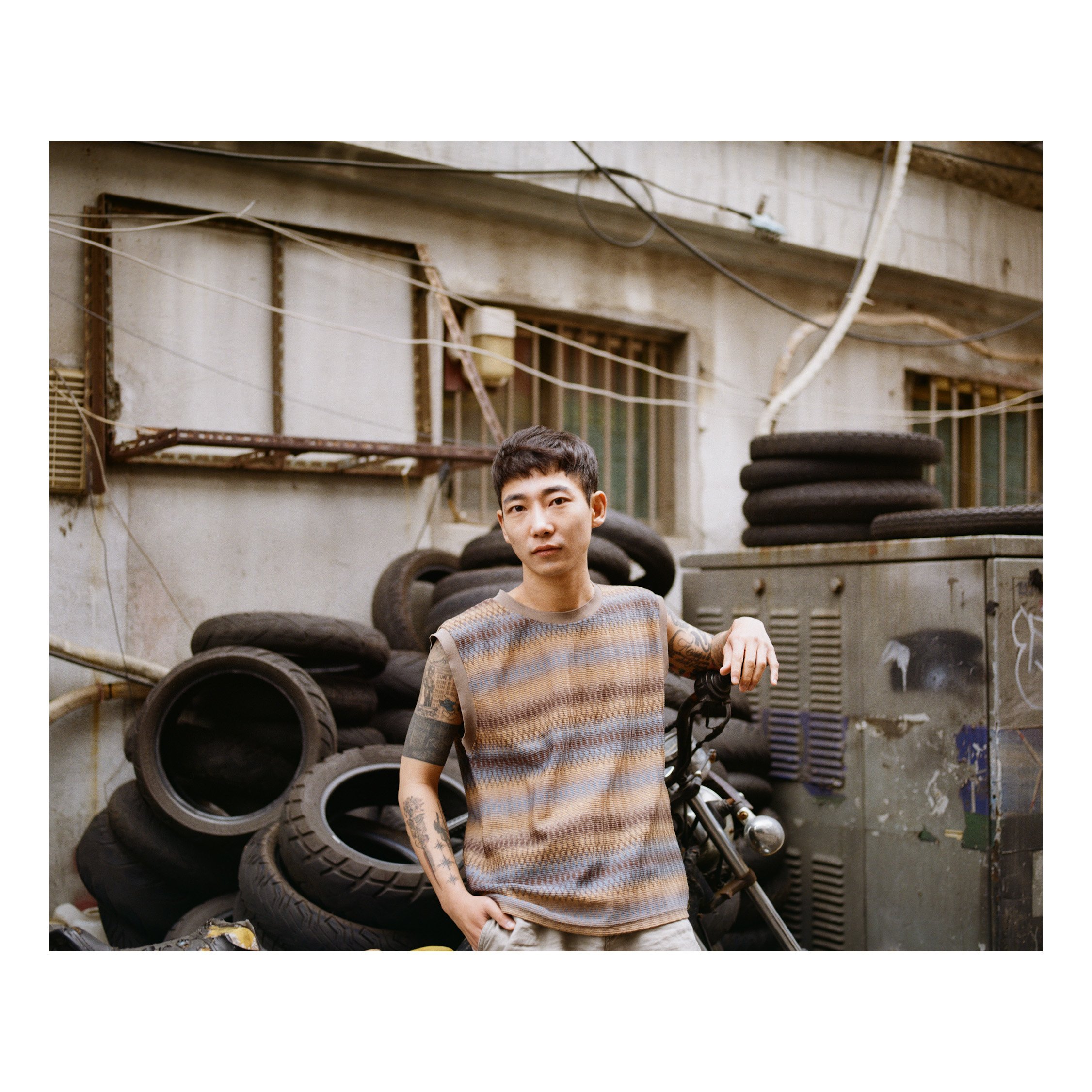 Young man with tattoos on arms standing next to a motorcycle in front of a wall with stacked tires and a metal utility box.