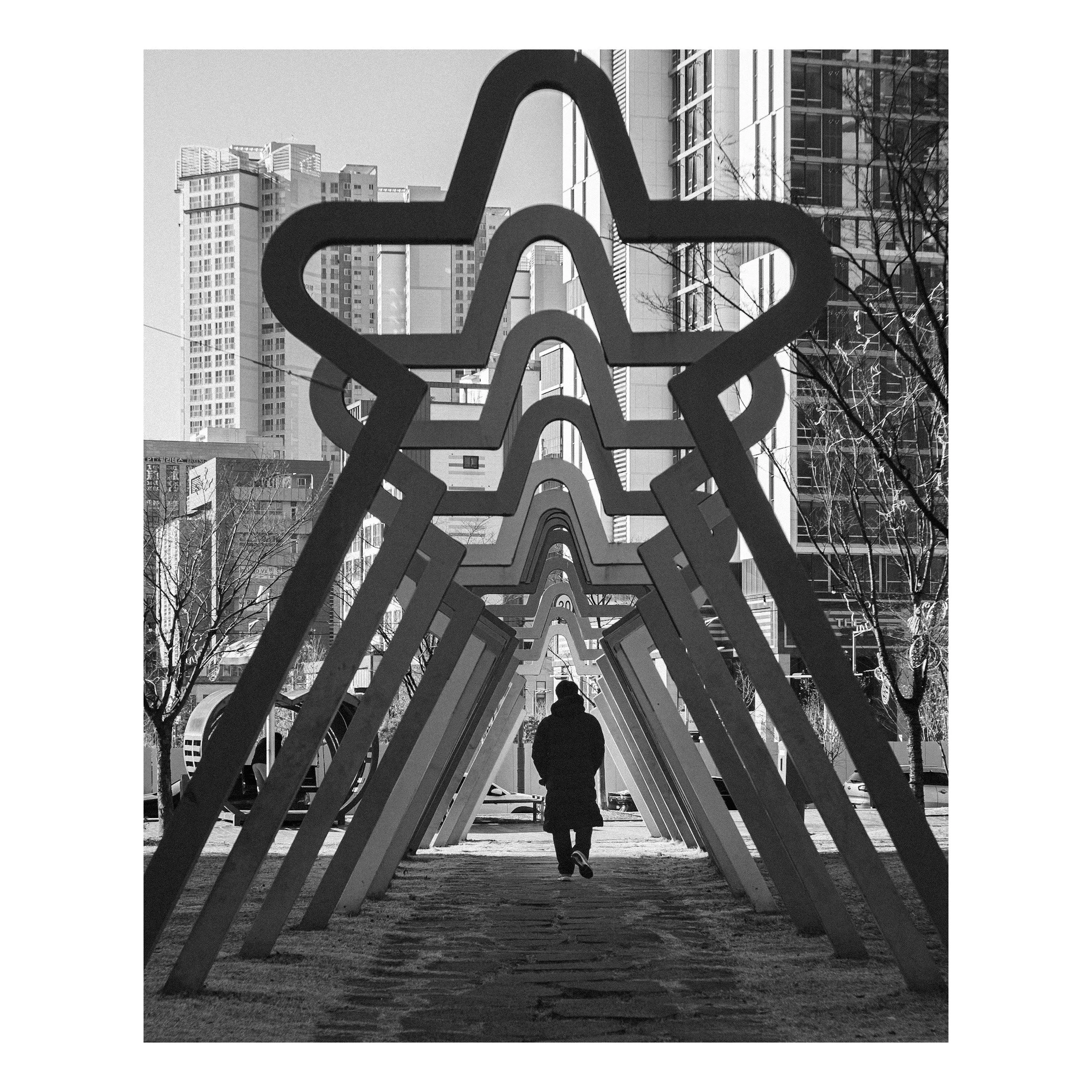 A person walking through an urban park with large star-shaped sculptures arranged in a line, with tall buildings in the background.
