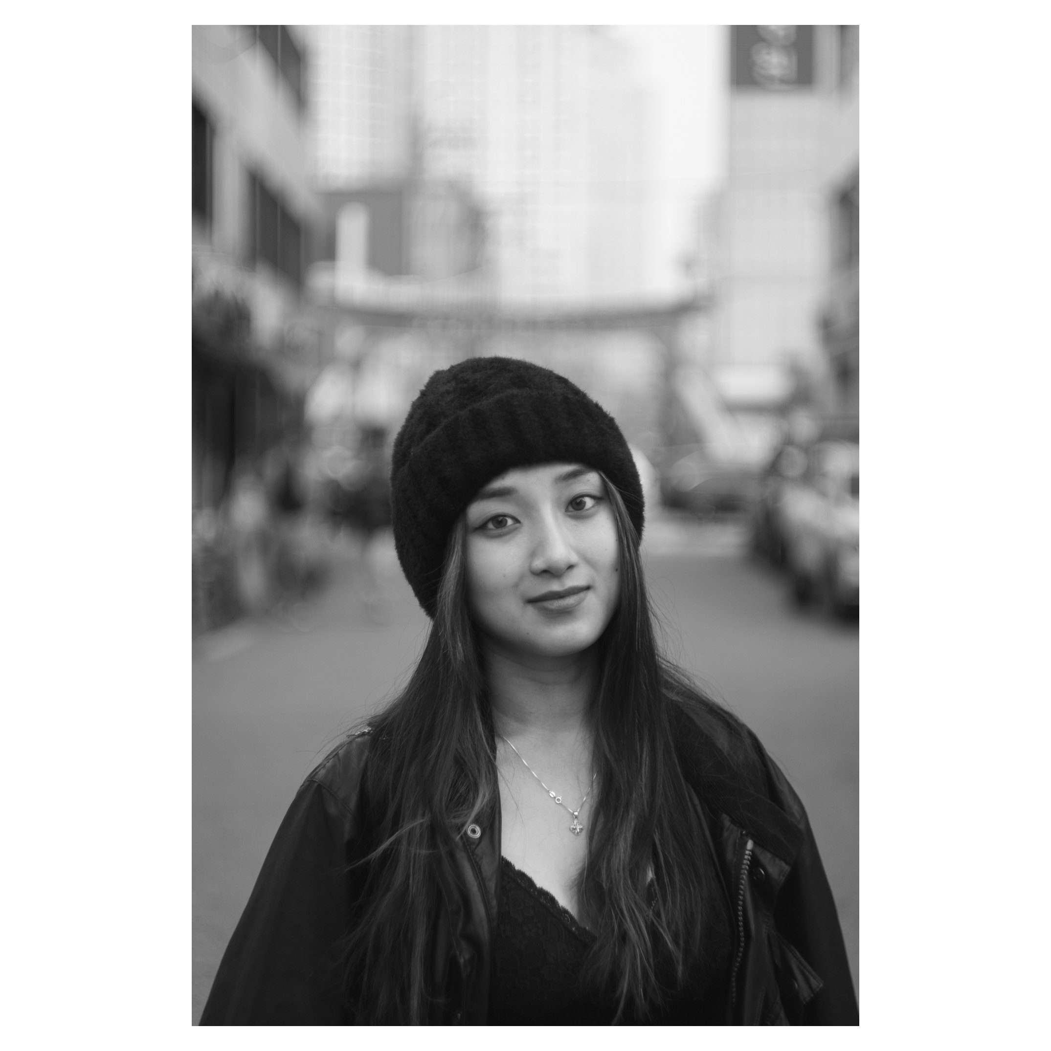Black and white photo of a young woman with long hair wearing a knit hat, outdoors on a city street with buildings and cars in the background.