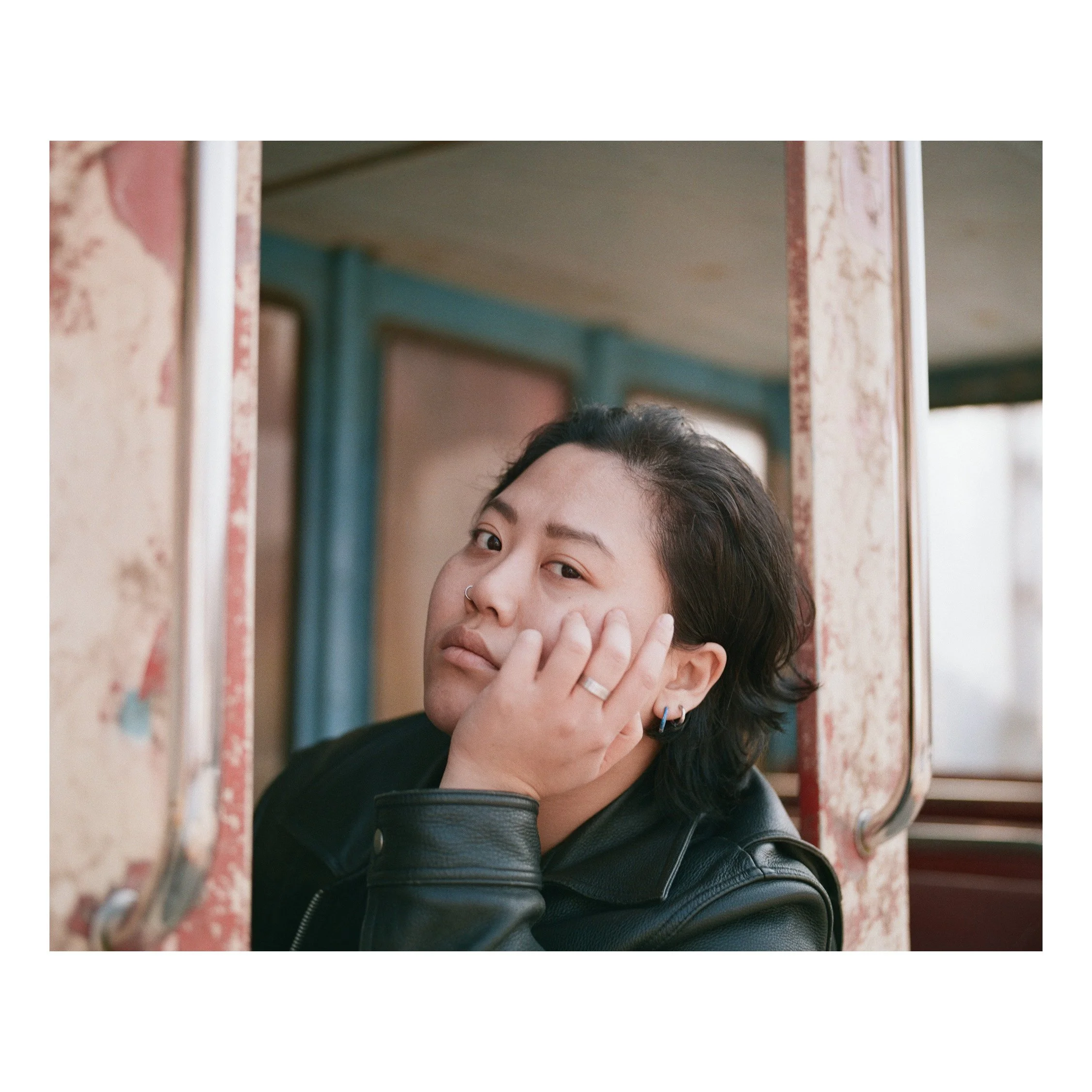 A woman with short dark hair, wearing earrings and a nose ring, looks at the camera while resting her head on her hand, seated inside an old, weathered train carriage.