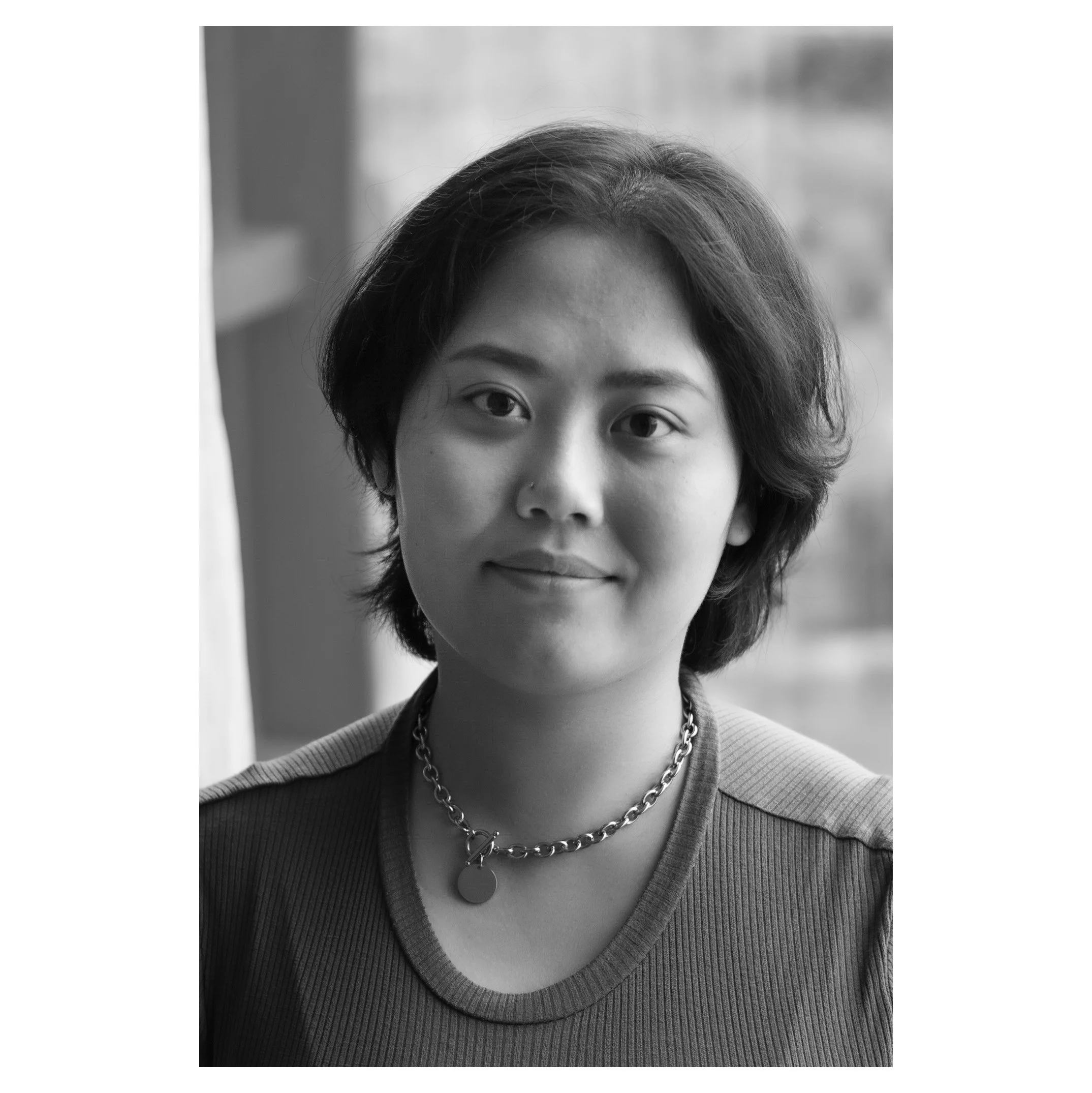 Black and white portrait of a woman with short, wavy hair, wearing a round-neck top and a chain necklace with a pendant, looking at the camera.