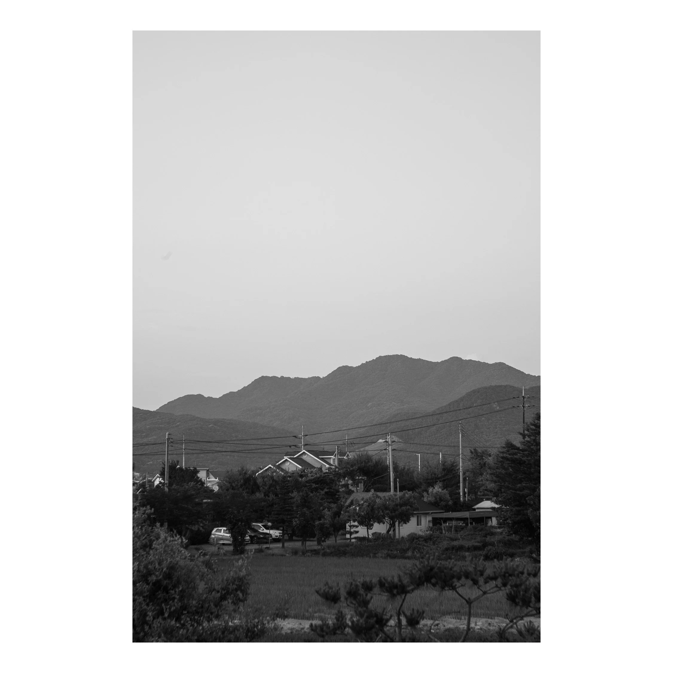 Scenic view of a rural neighborhood with houses and trees in the foreground, power lines, and mountains in the background under a clear sky. Black and white photo.