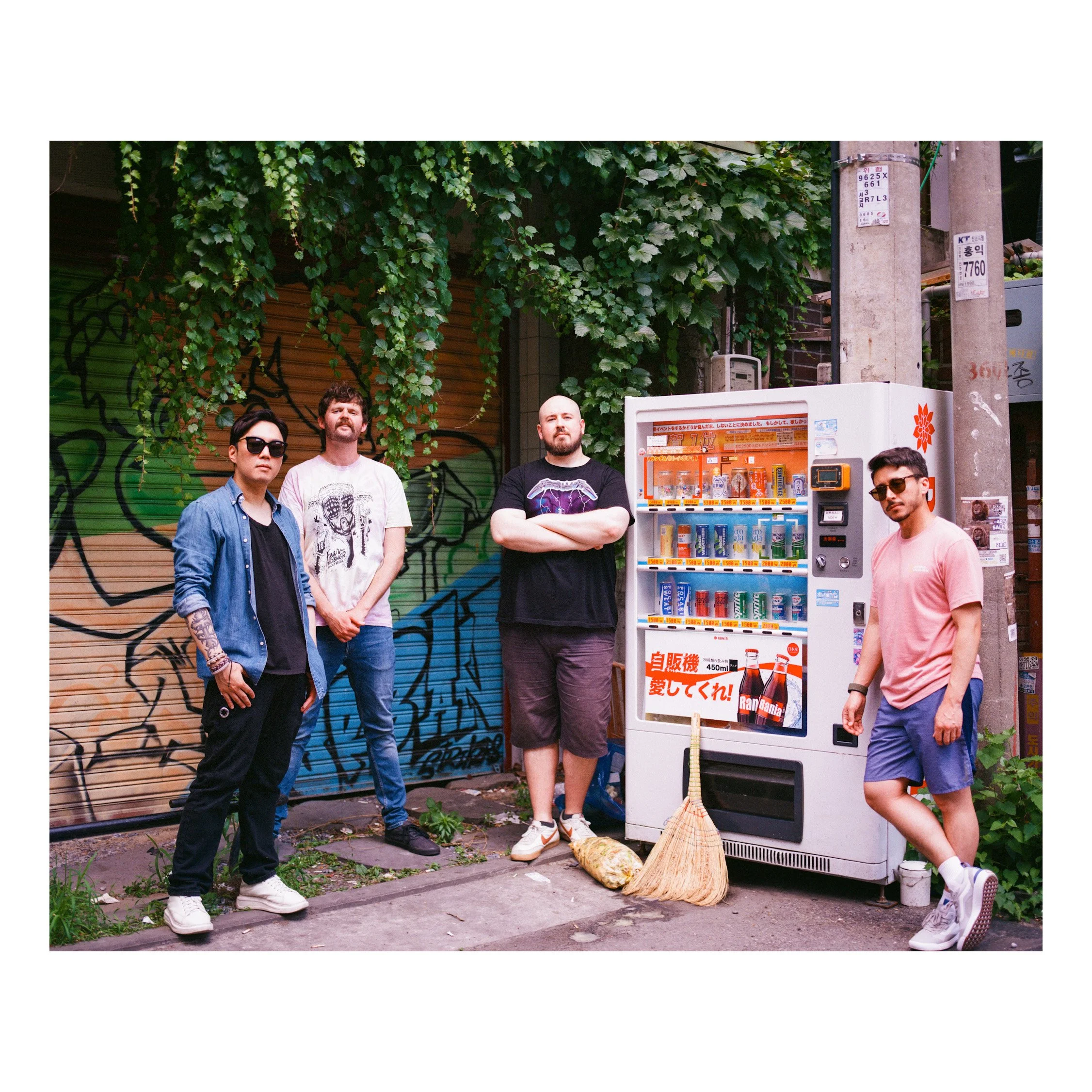 Four men standing next to a vending machine on a city street with graffiti and greenery in the background.