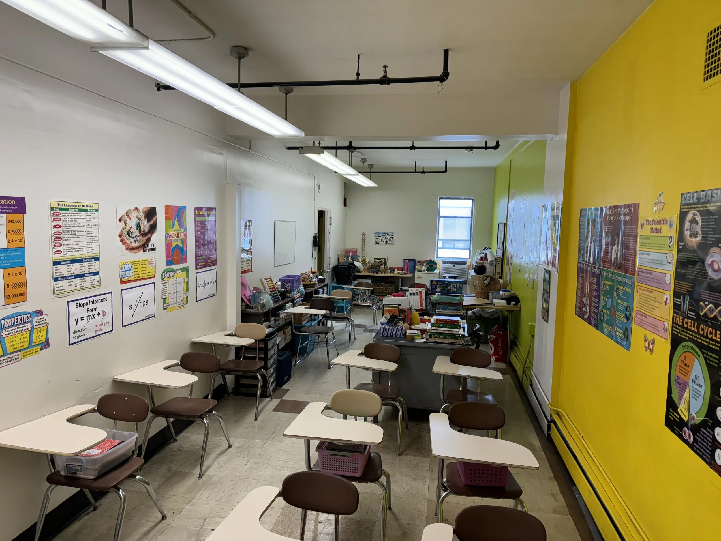 Empty classroom with desks and chairs, yellow and white walls, educational posters, and supplies on desks and shelves.