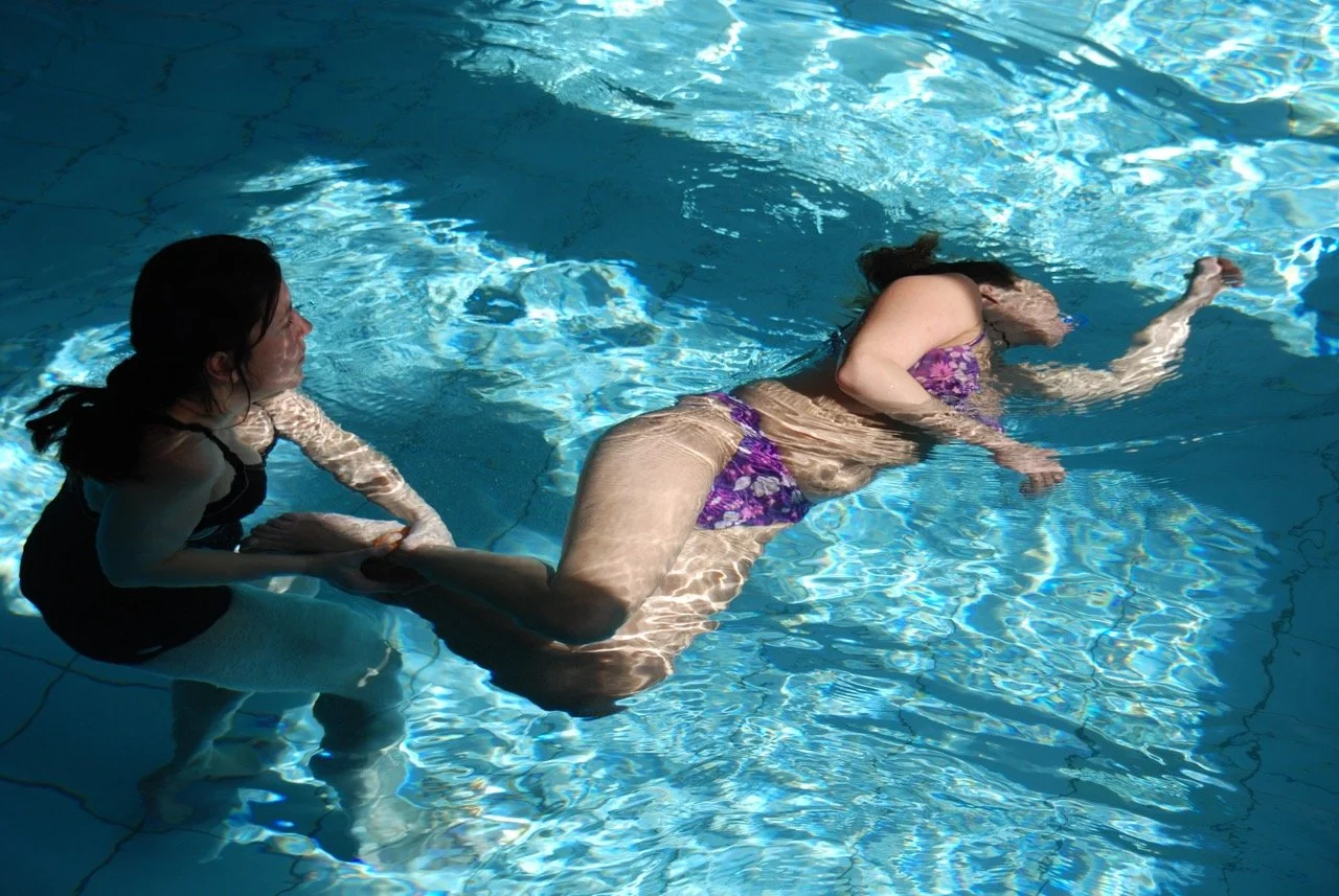 Zwei Personen im klaren Wasser. Die eine Person wendet Floating Therapie an. Sie hält die zweite Person dabei an den fußgelenken und schwingt ihren Körper sanft durchs Wasser. Die Person floatet dabei seitlich und ist unter Wasser.