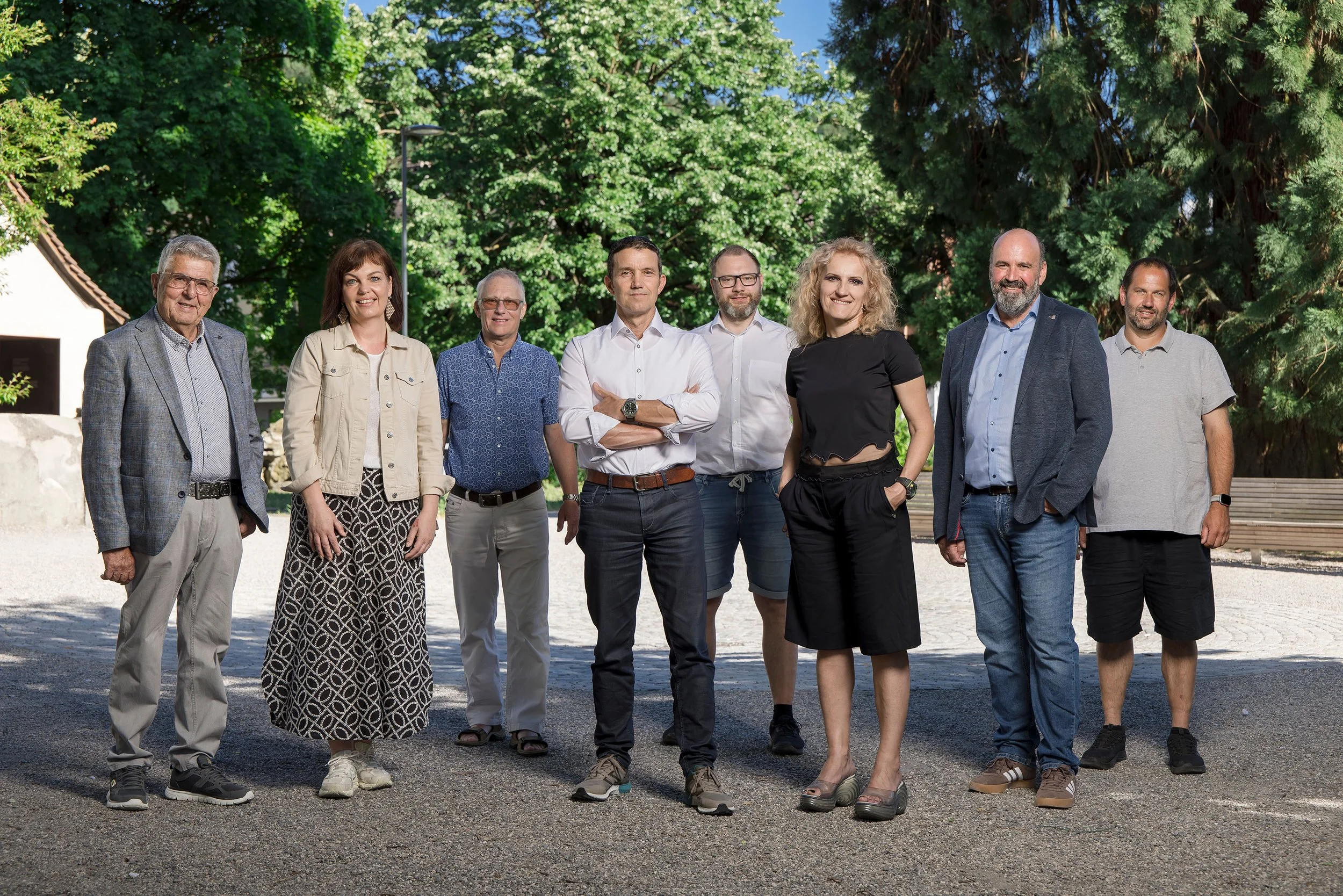 Eine Gruppe von acht Personen der Volksparte Rankweil steht im Freien auf einem gepflasterten Platz, umgeben von grünen Bäumen.