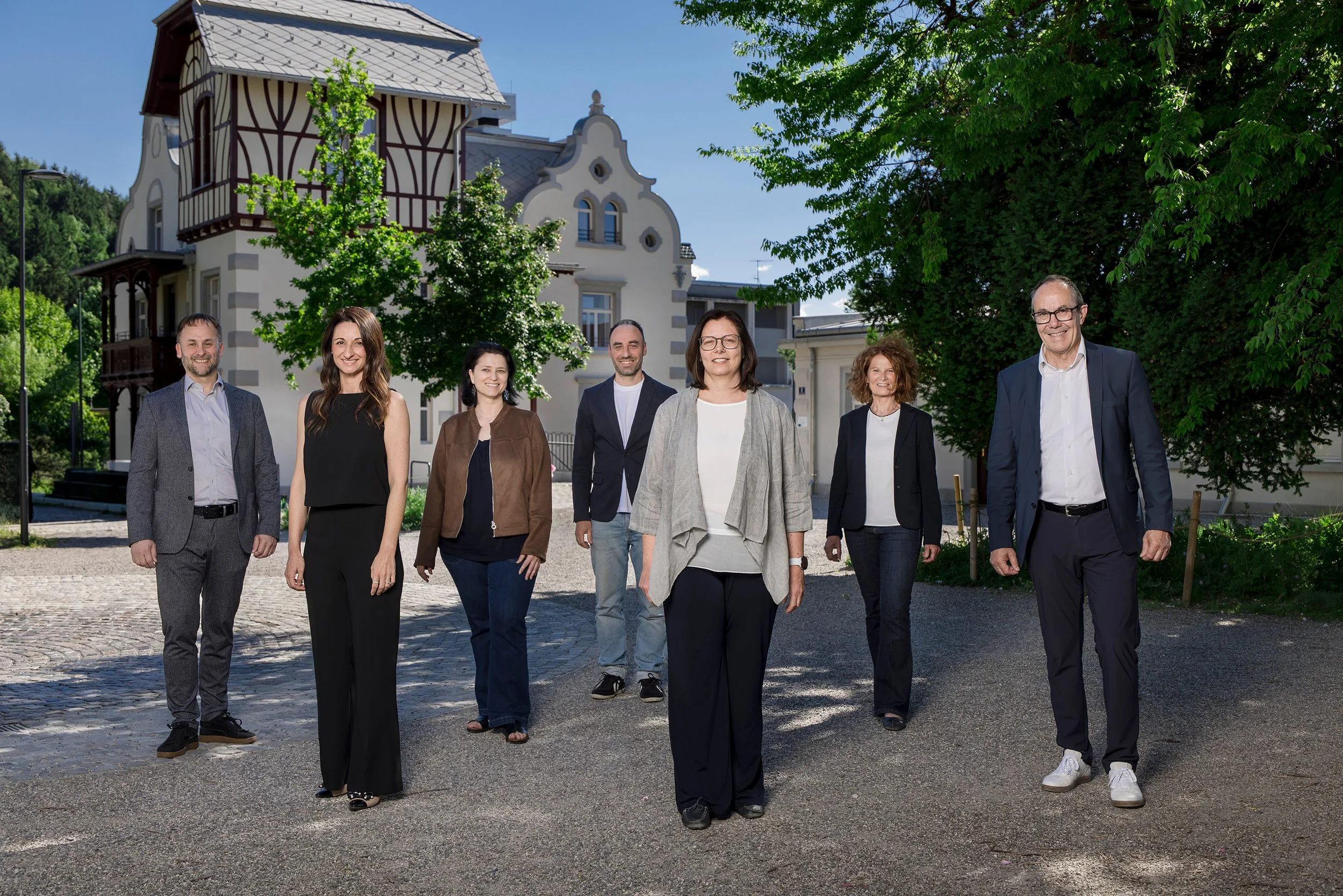 Gruppe von sieben Personen, Männer und Frauen, steht auf einer gepflasterten Straße vor der Häusle Villa in Rankweil umgeben von grünen Bäumen bei Sonnenschein.