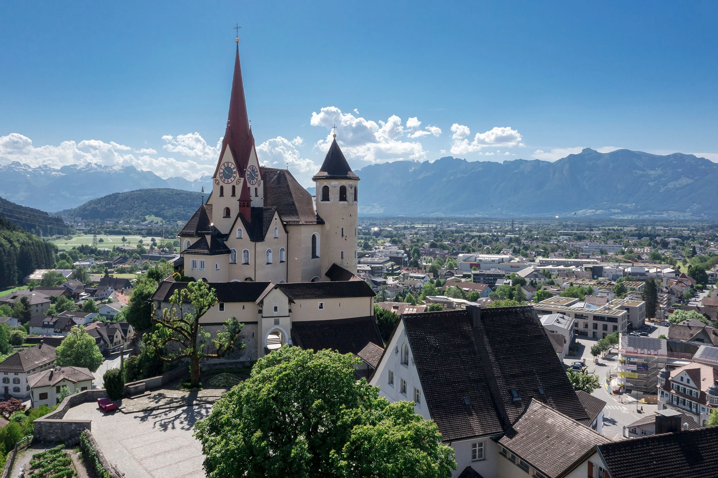 Ein Stadtbild von der Basilika in Rankweil, umgeben von Wohnhäusern und grünen Bäumen, vor einer Bergkulisse unter einem blauen Himmel mit Wolken.