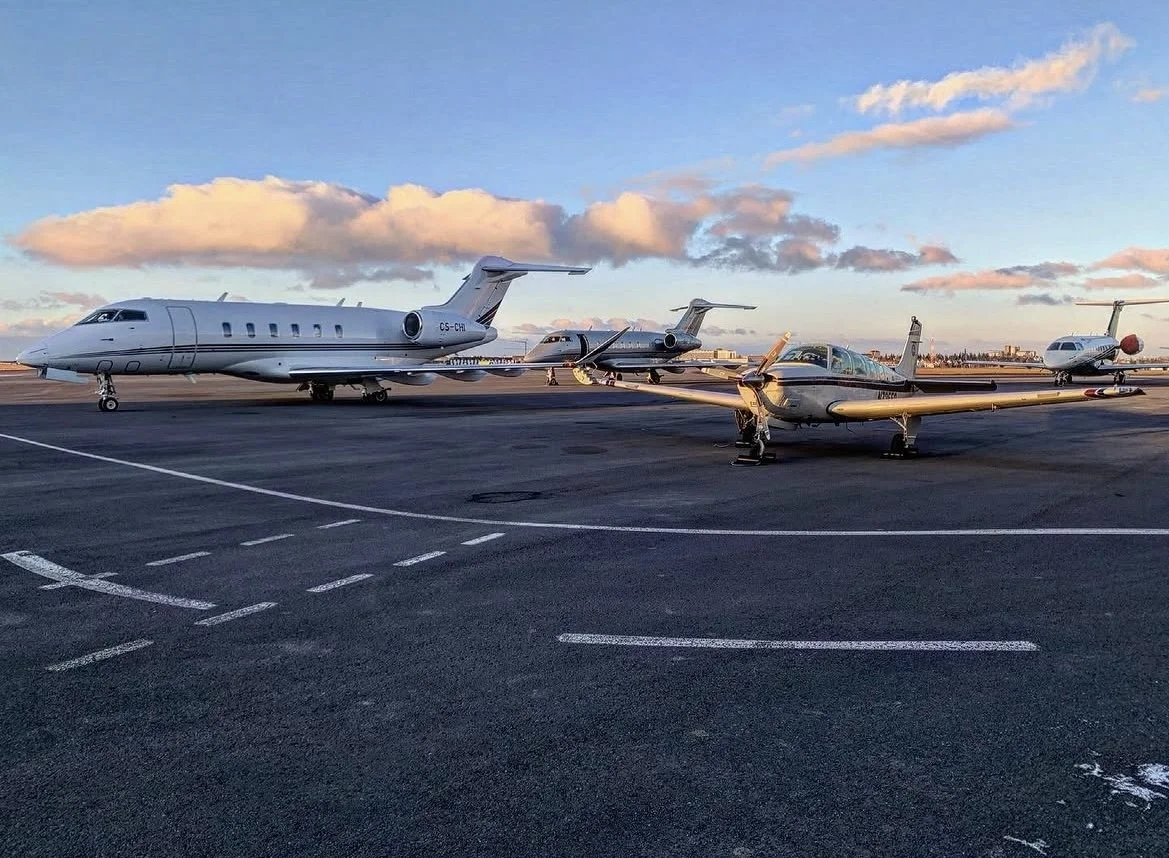 Multiple private jets parked on an airport tarmac during sunset with clouds in the sky.