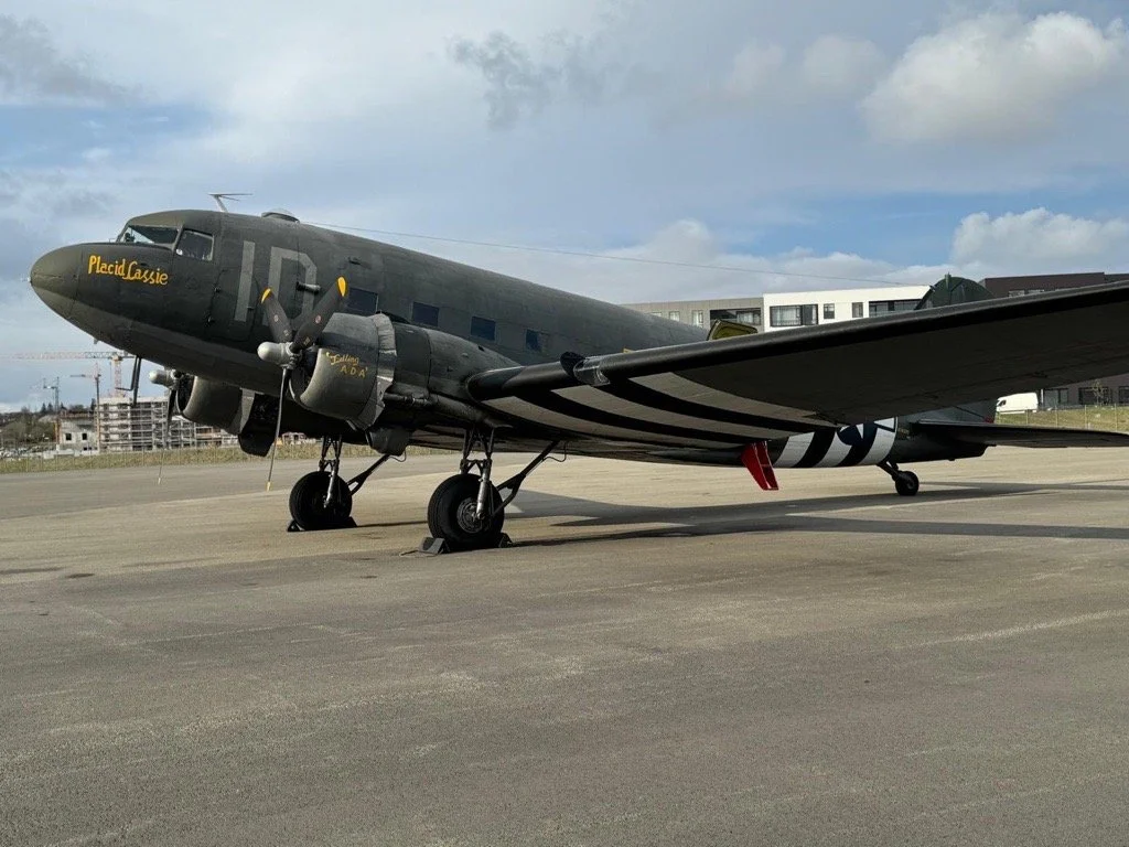 A vintage military aircraft on the tarmac with black and white stripes, a dark gray body, and the name 'Placid Cassie' written on the nose.