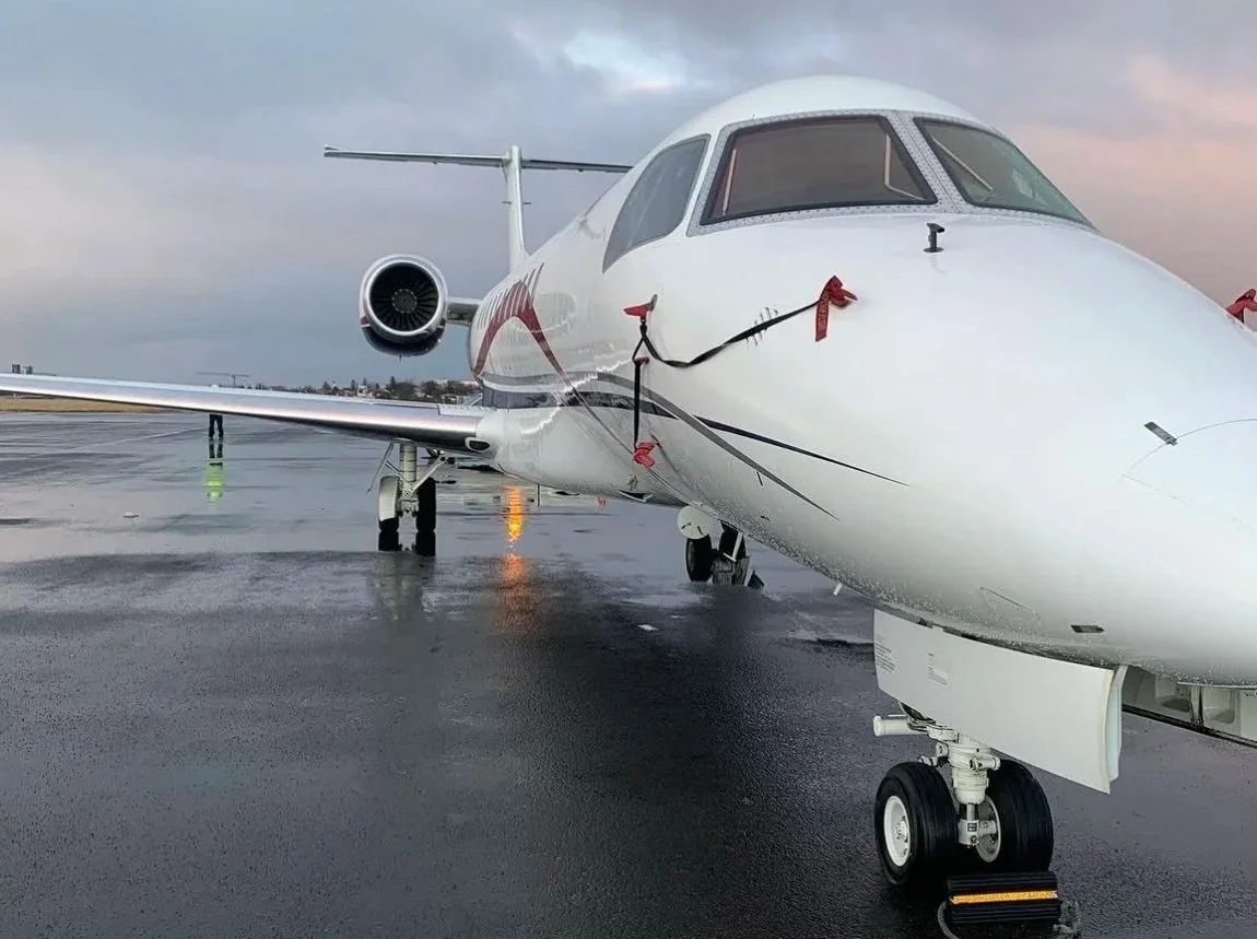 Private jet on wet tarmac with tied-down straps, overcast sky in the background.