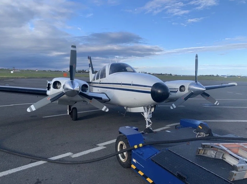 A twin-engine propeller airplane parked on the tarmac at an airport, connected to ground support equipment, with a blue sky and clouds overhead.