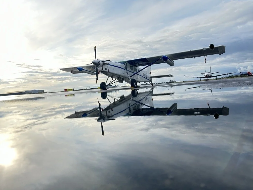 Small twin-engine airplane parked on a tarmac, with its reflection visible on a wet surface, under a cloudy sky.