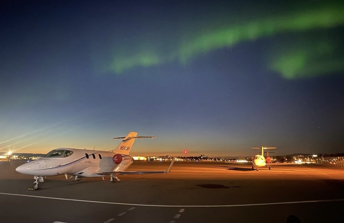 Two white private jets parked on an airport tarmac at dusk, with the Northern Lights visible in the night sky above.
