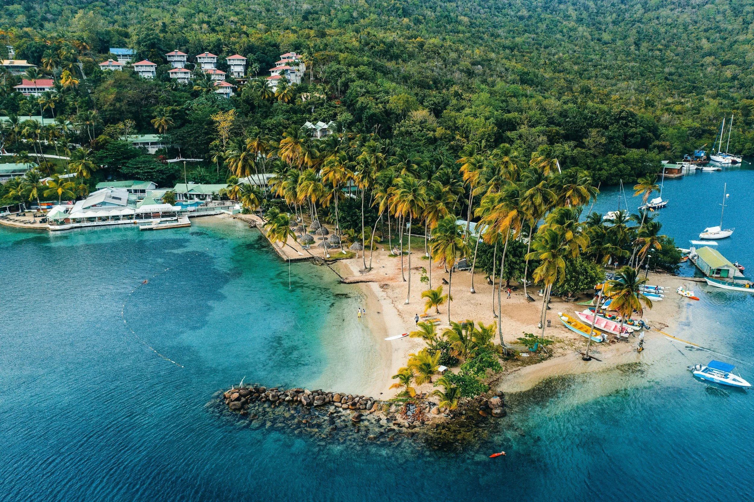 Aerial view of a tropical beach with palm trees, clear blue water, boats, and a resort with buildings on a hillside.