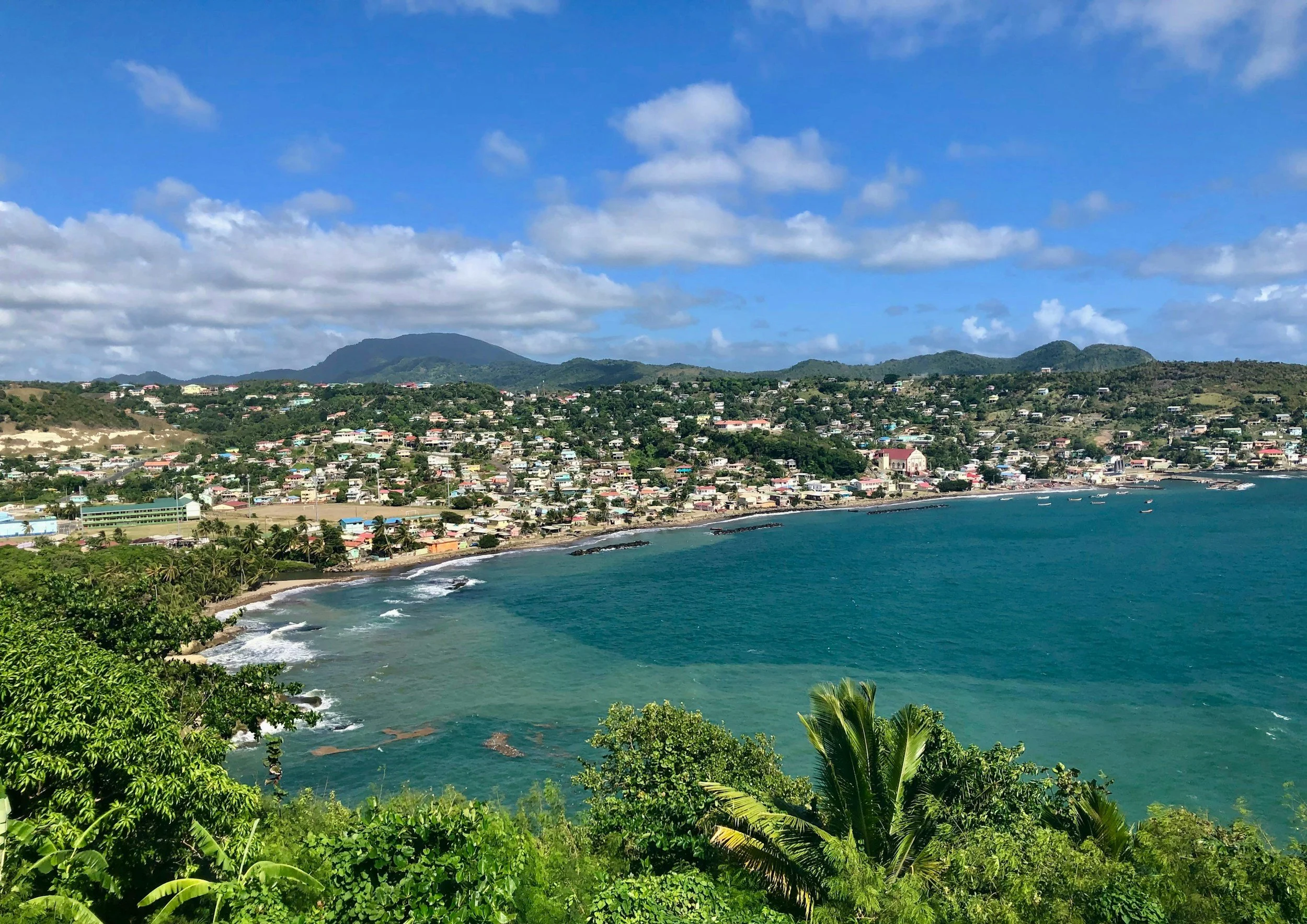 A coastal town surrounded by green hills and mountains under a blue sky with fluffy clouds, with boats near the shoreline and lush tropical vegetation in the foreground.