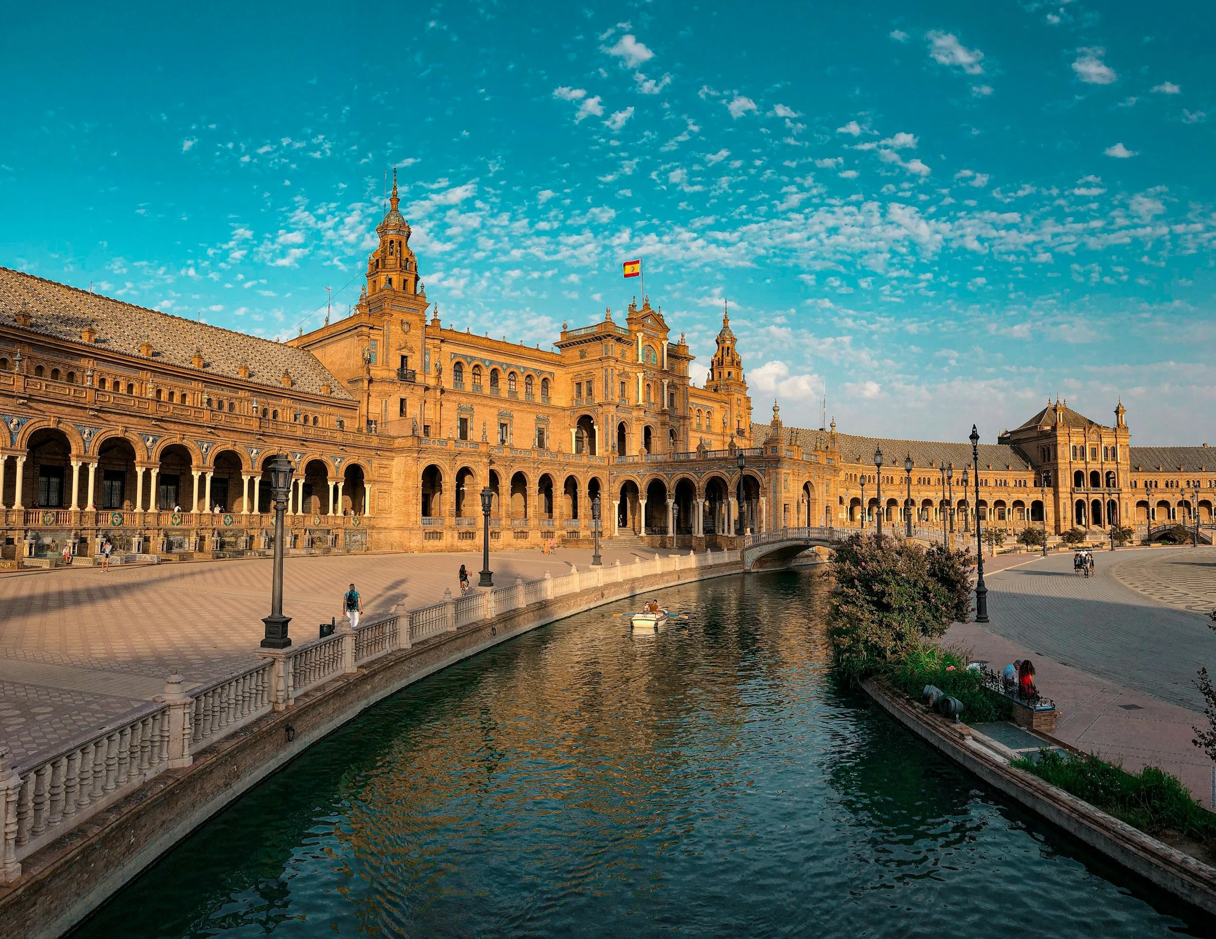Seville Spain Alcázar illuminated by sunlight, with a waterway and people walking nearby, under a blue sky with scattered clouds.