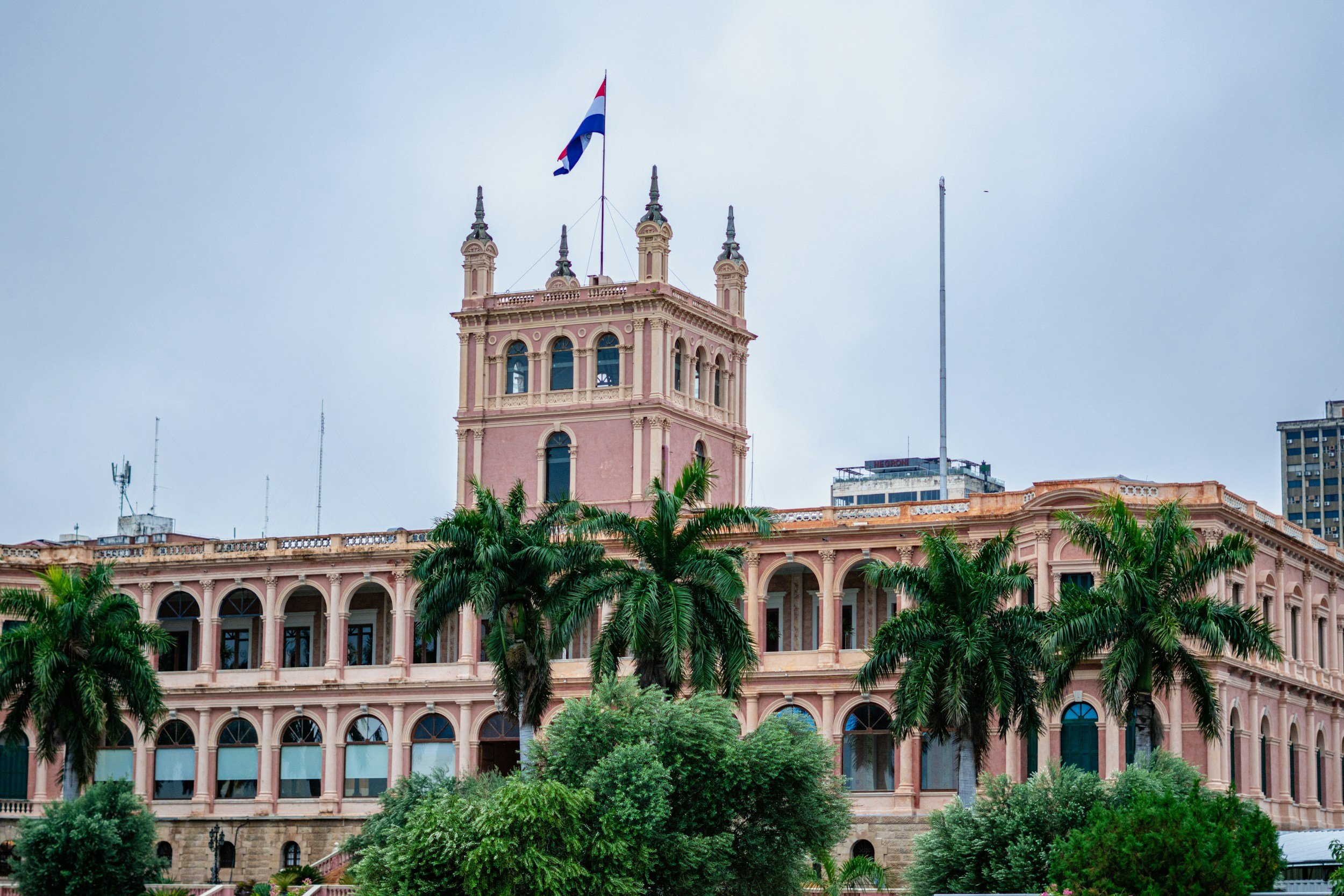 Pink historic building with a tower flying a flag, palm trees and lush green shrubs in front, overcast sky in the background.