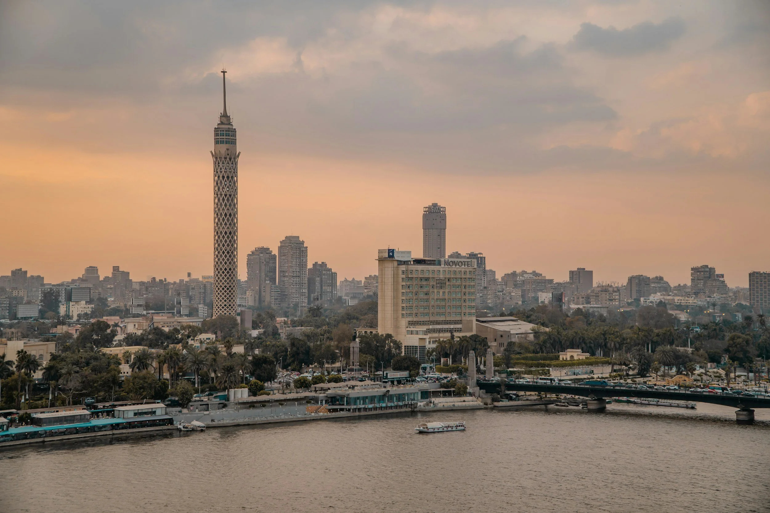City skyline with a tall, lattice-patterned tower near a river, showing buildings, trees, a bridge, and boats at sunset.