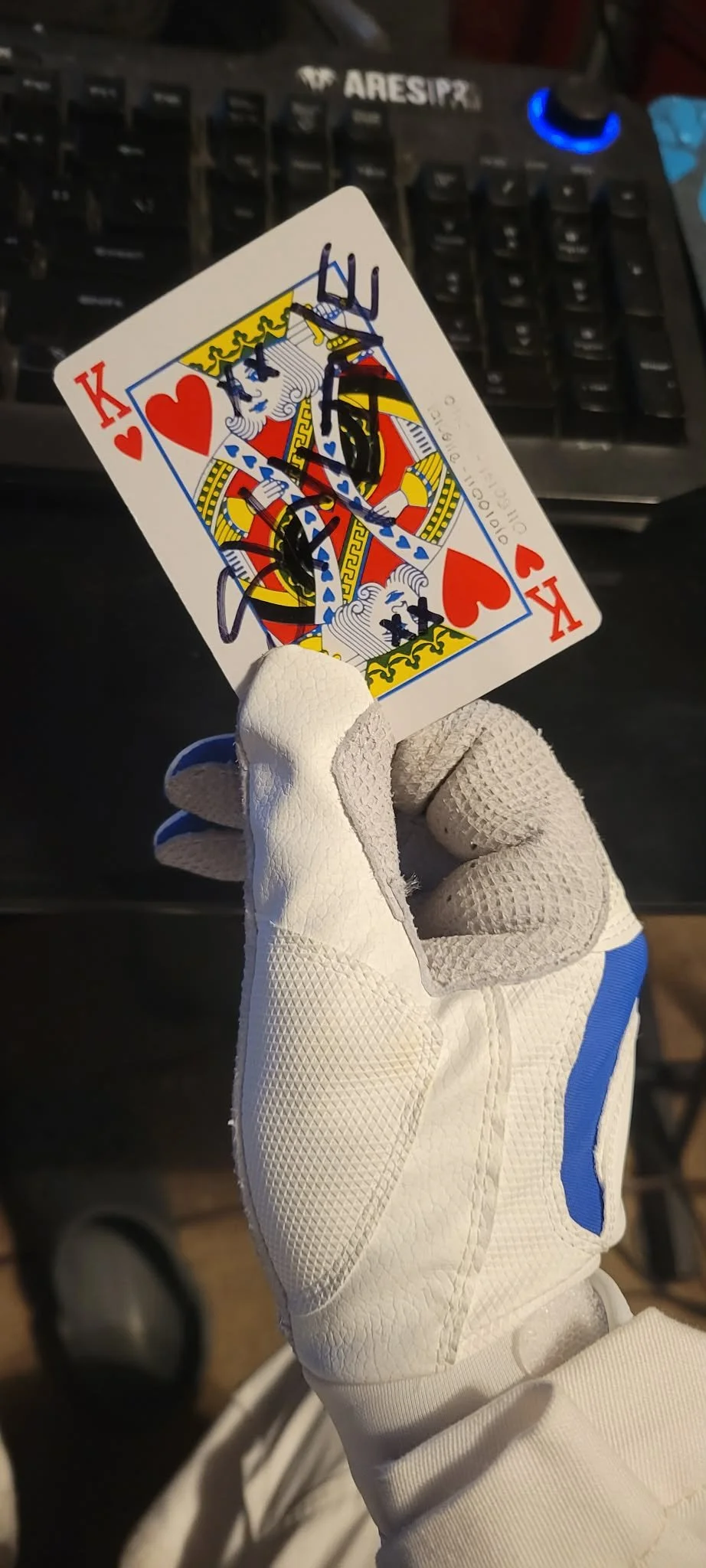 A hand wearing a white golf glove holding a King of Hearts playing card with a black signature on it. The background includes a black computer keyboard.